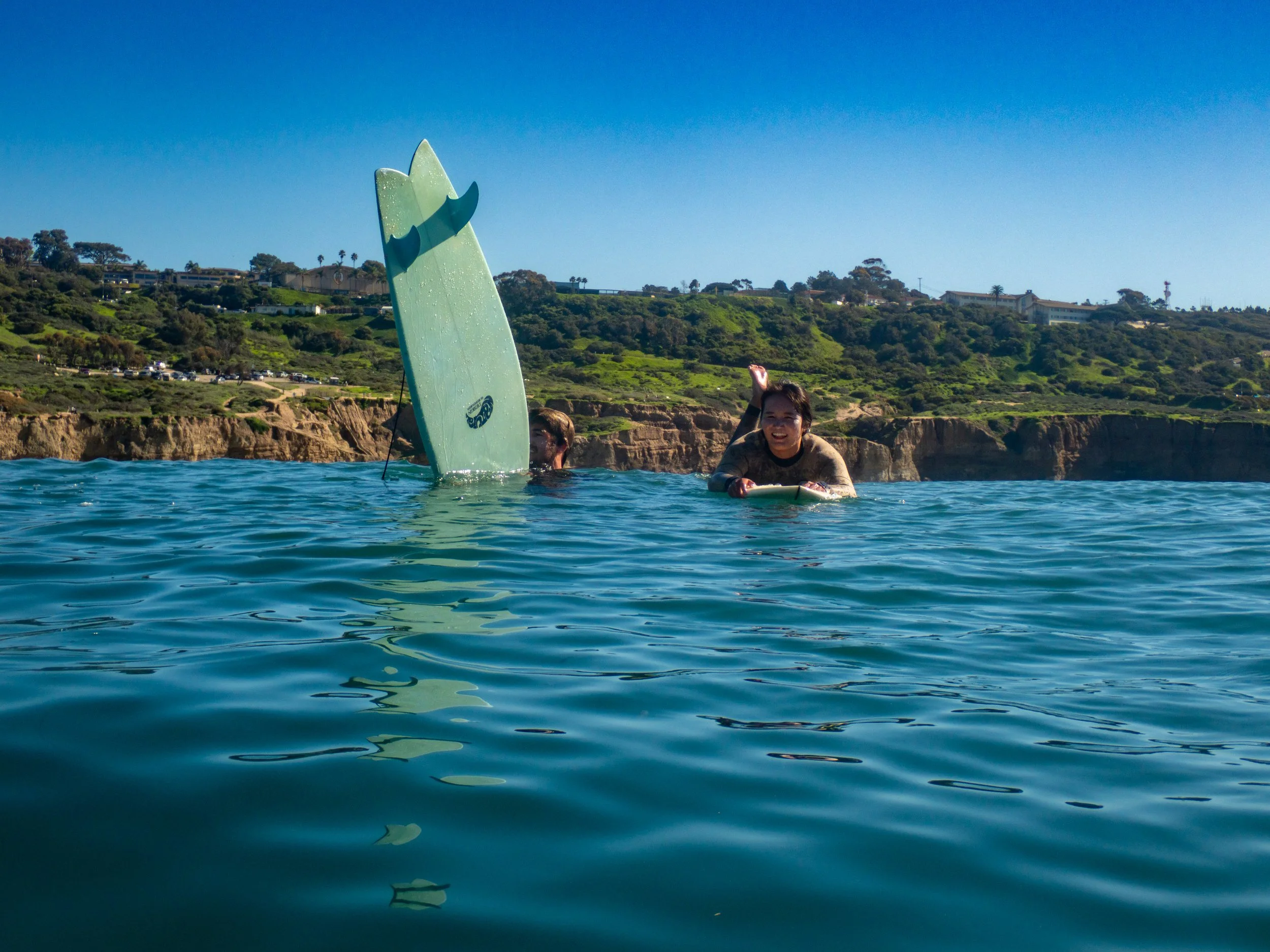 Two people in the ocean, one with a partially submerged surfboard and the other holding a surfboard, near a rocky coastline with green hills and houses in the background during sunny weather.