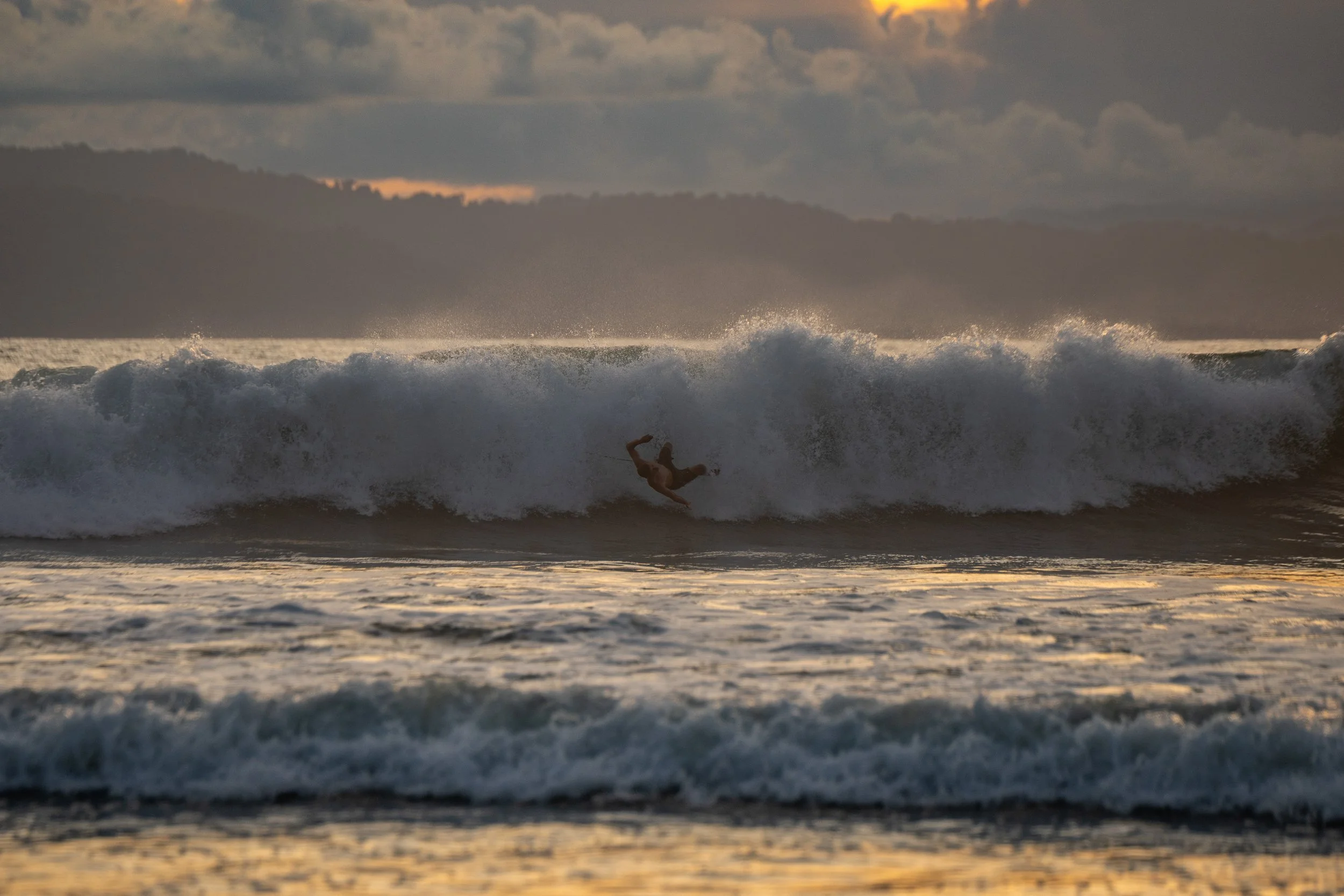 A person falling off a surfboard in the ocean during sunset with waves crashing and clouds in the sky.