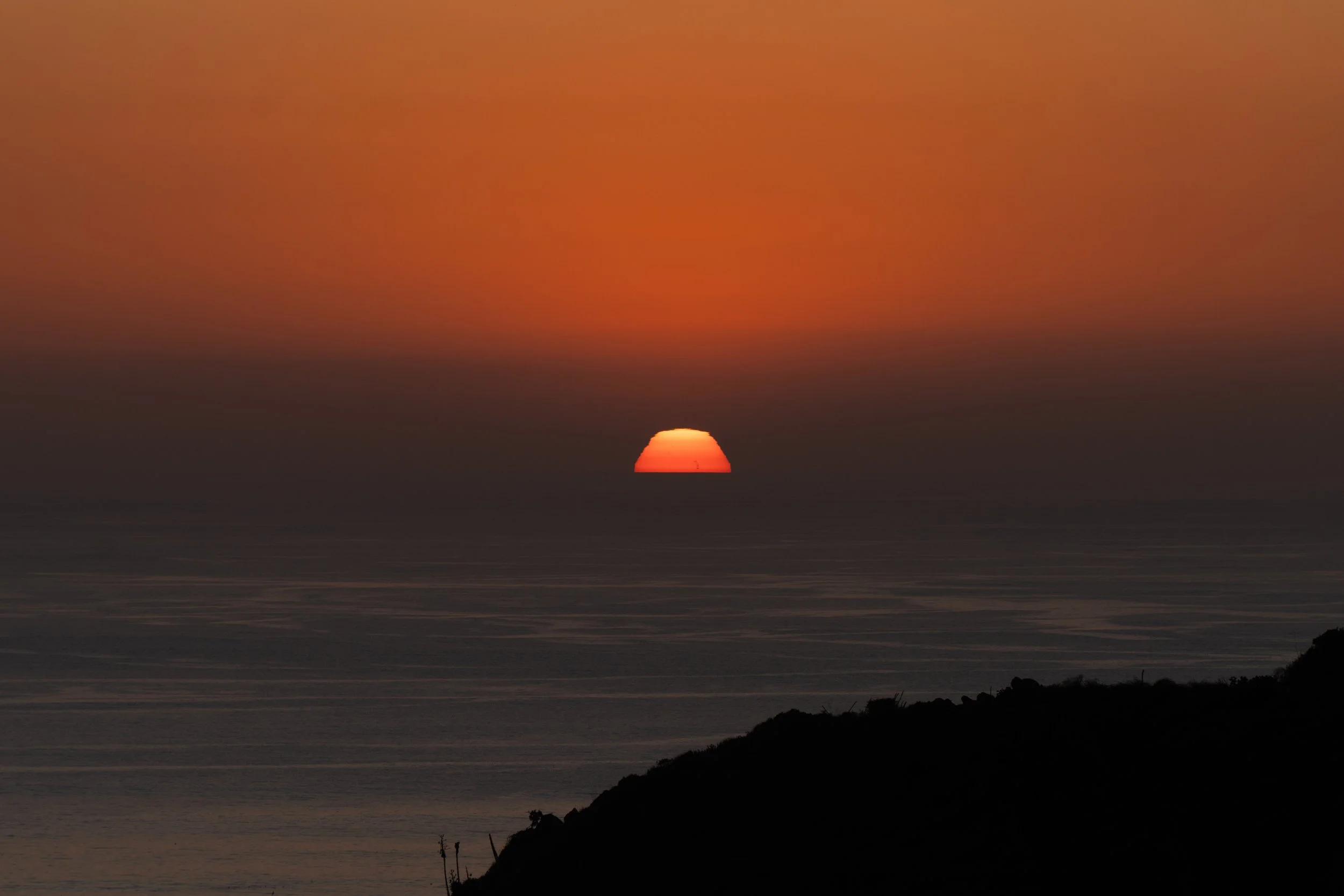 A sunset over the ocean with the sun partially below the horizon, casting an orange glow across the sky and water, with a silhouette of a hillside in the foreground.