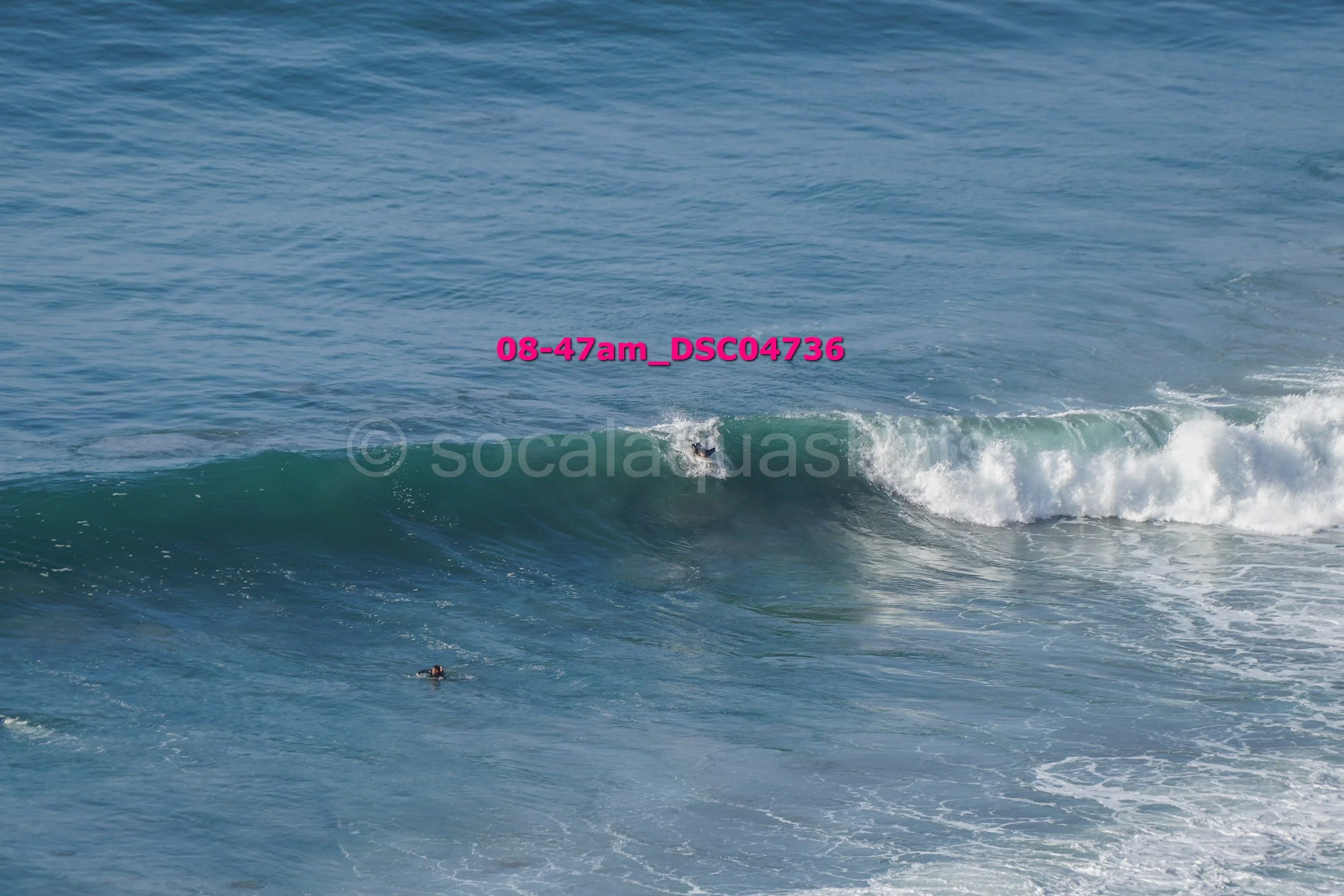 A person surfing on a small wave in the ocean, with another swimmer visible nearby.