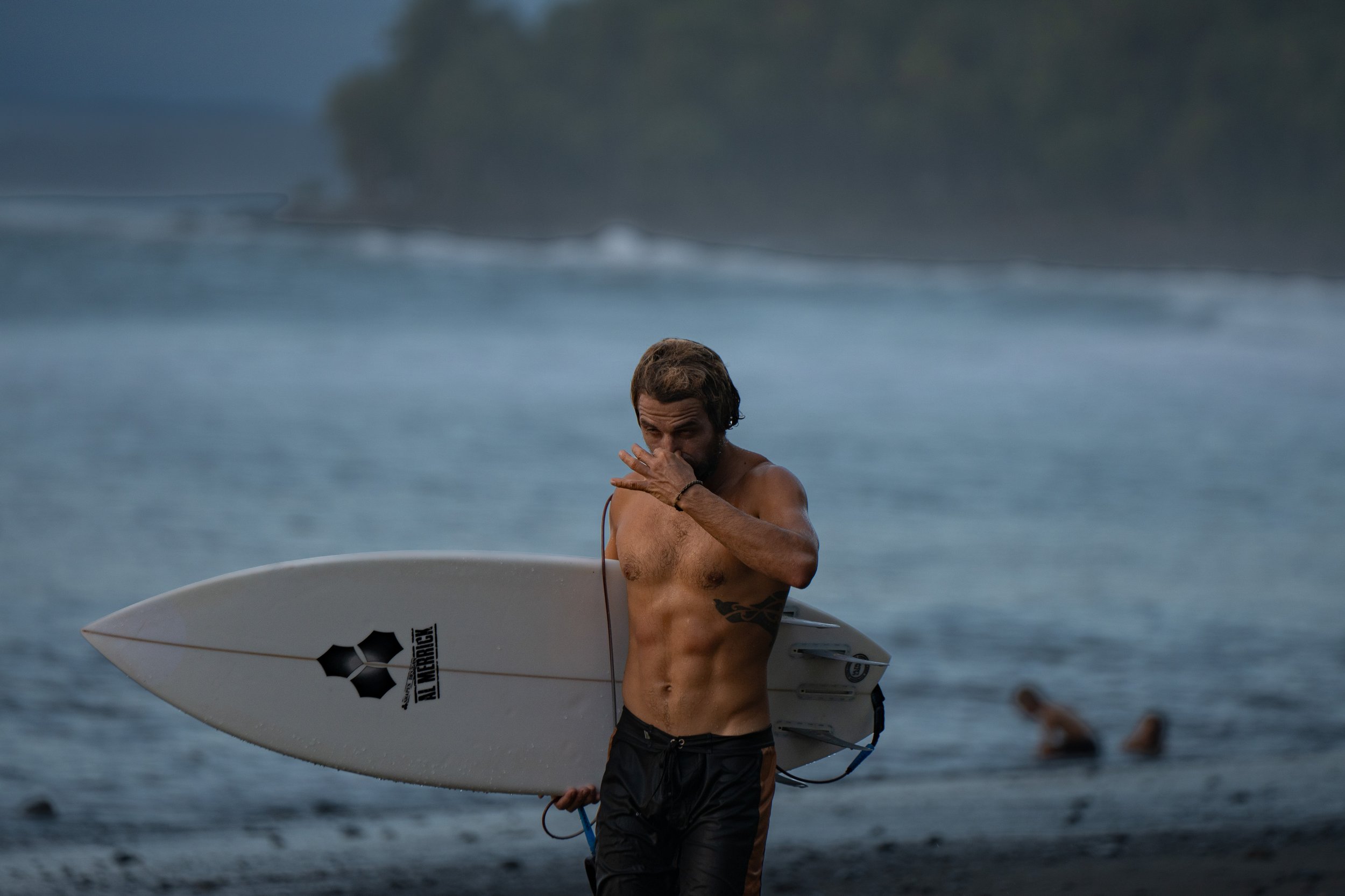 A man with wet hair and a beard holding a surfboard with a black logo on the side, standing in shallow water at the beach during the daytime.