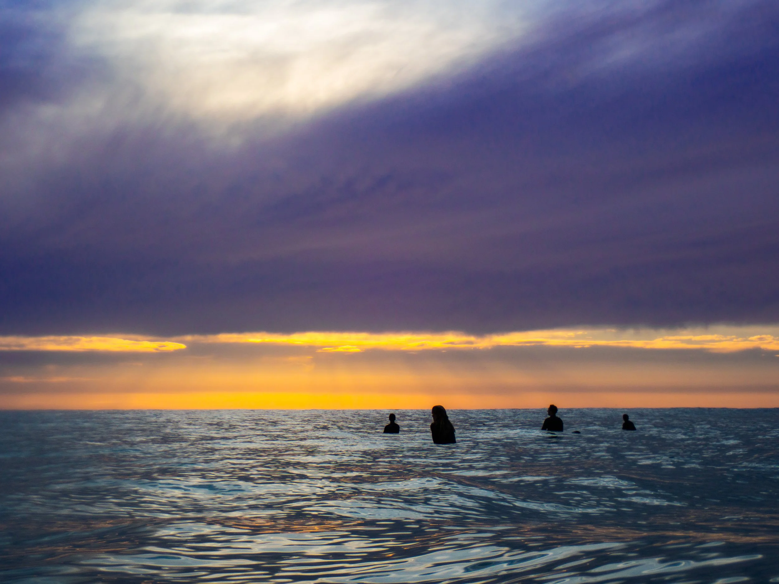 People standing in the ocean water at sunset with dark clouds and a colorful sky in the background.