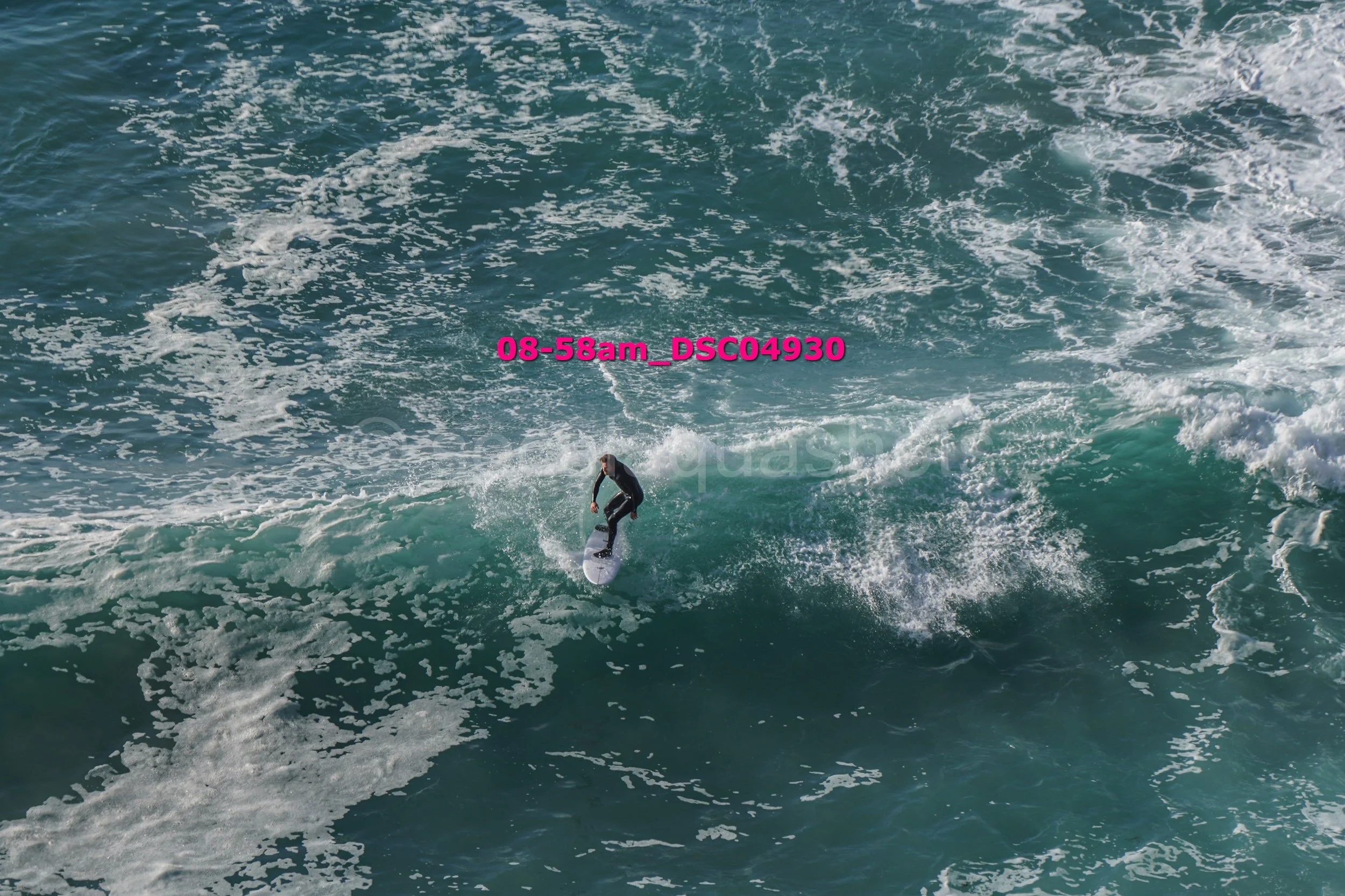 A person surfing on a large ocean wave with white foam, wearing a wetsuit under a bright blue sky.