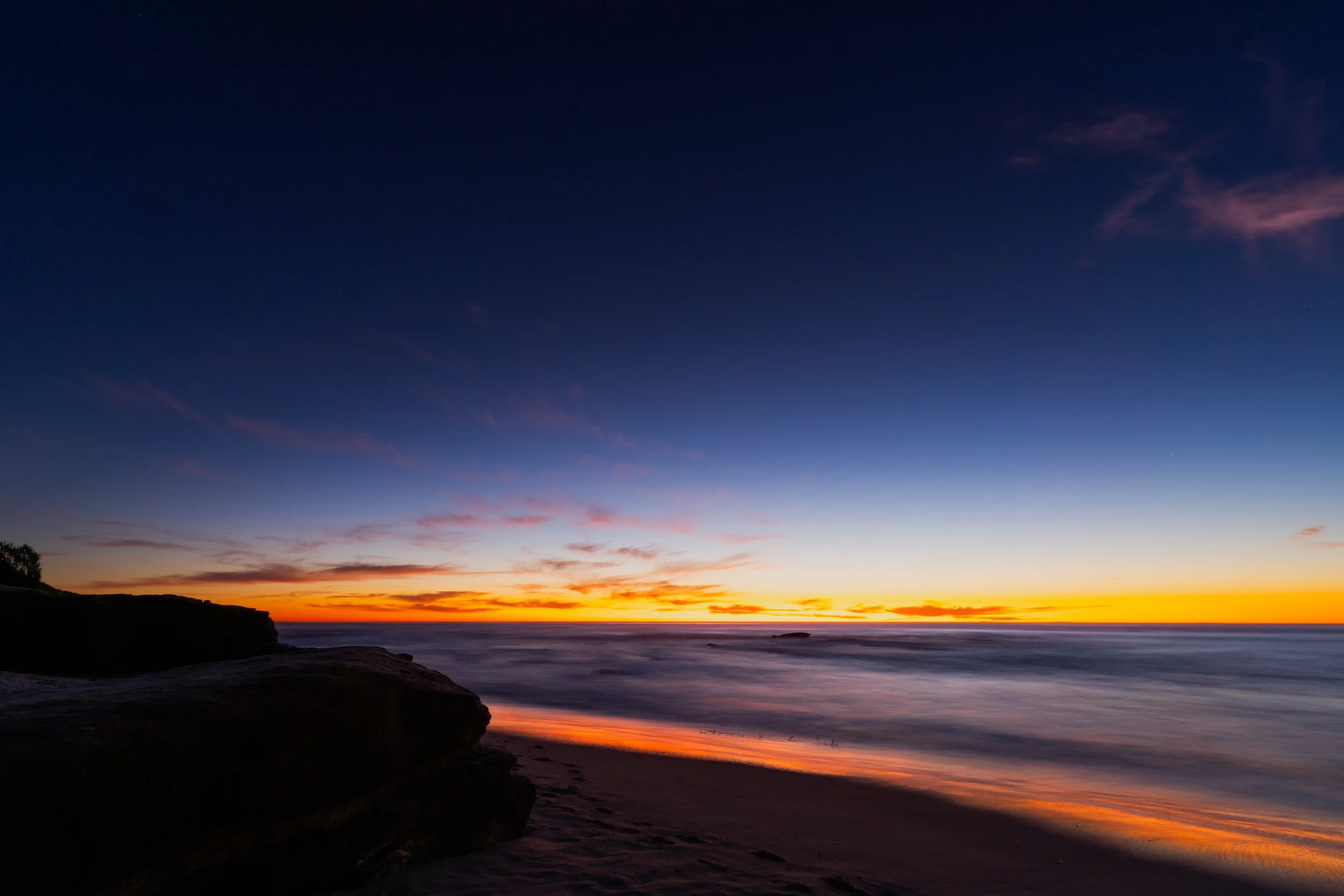 Sunset over the ocean with a clear sky transitioning from orange near the horizon to deep blue higher up, with rocks and a sandy beach in the foreground.