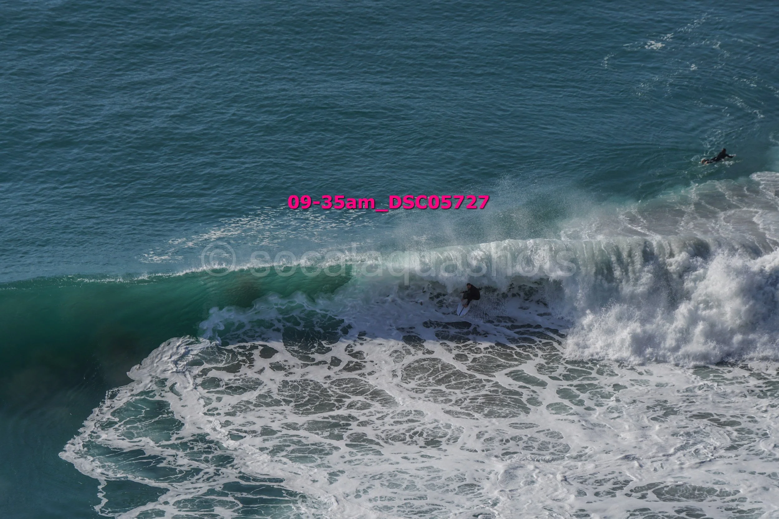 A person surfing on a wave in the ocean with another surfer in the distance. Water is blue and white, with foam and spray from the wave.