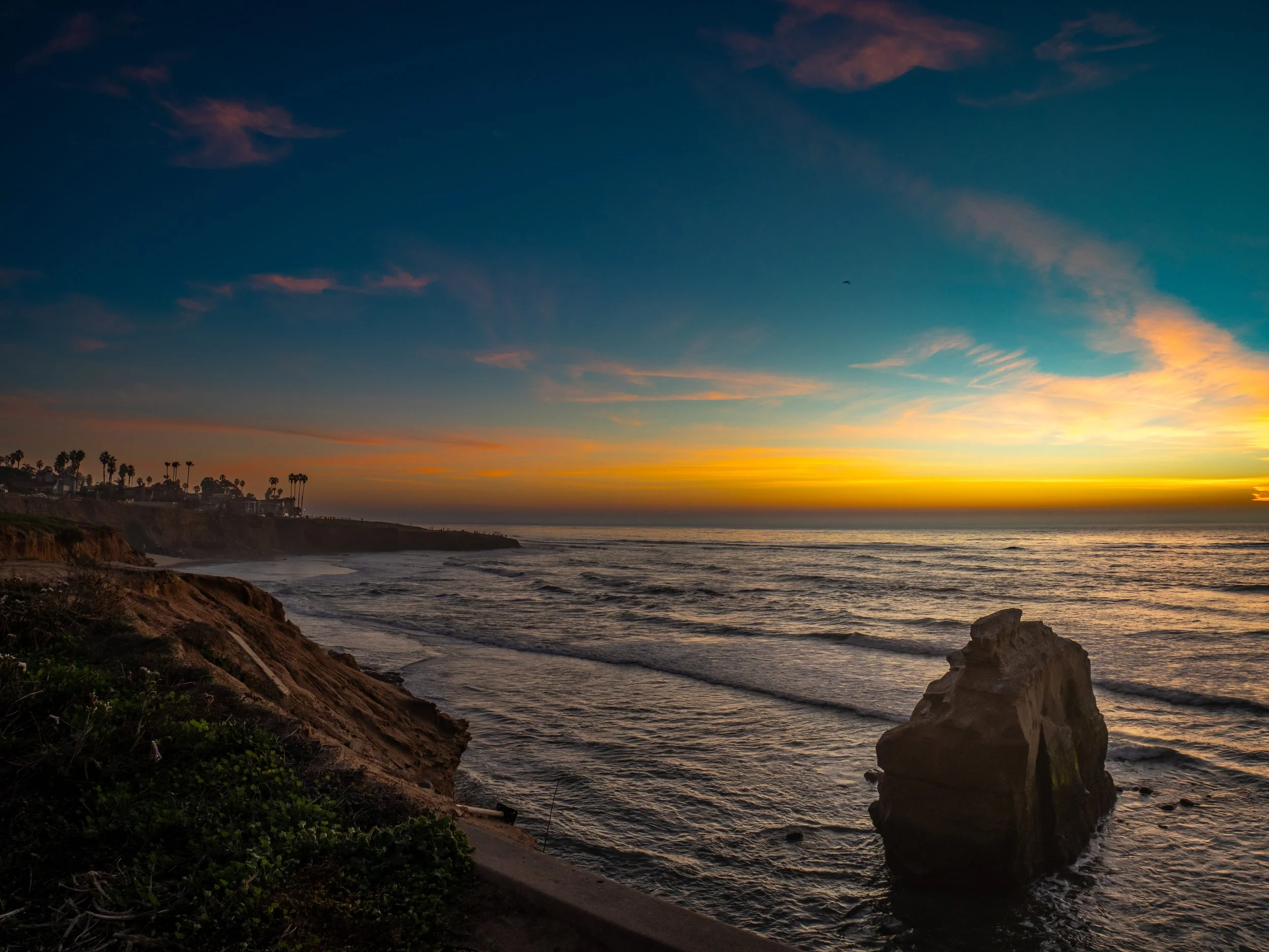 Sunset over the ocean with colorful sky, shoreline with cliffs and a large rock near the water, palm trees on the distant coast.