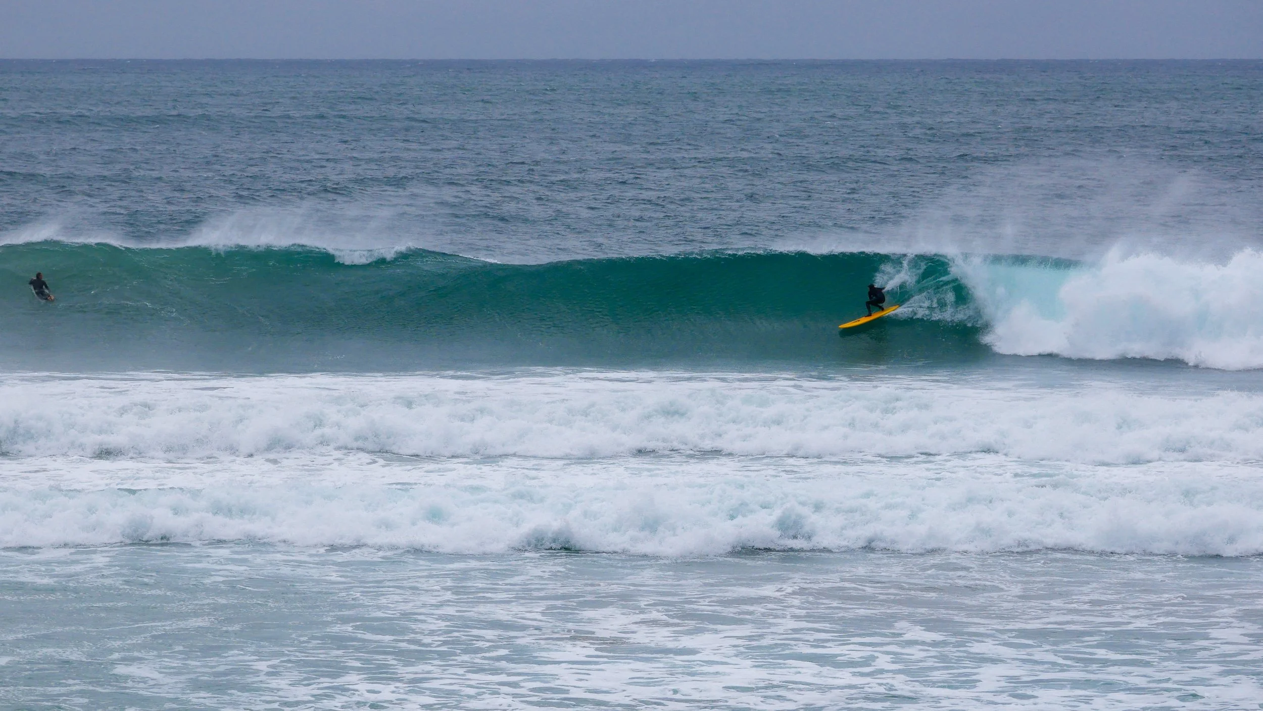 Surfer on a yellow board riding a wave in the ocean with another surfer in the background.