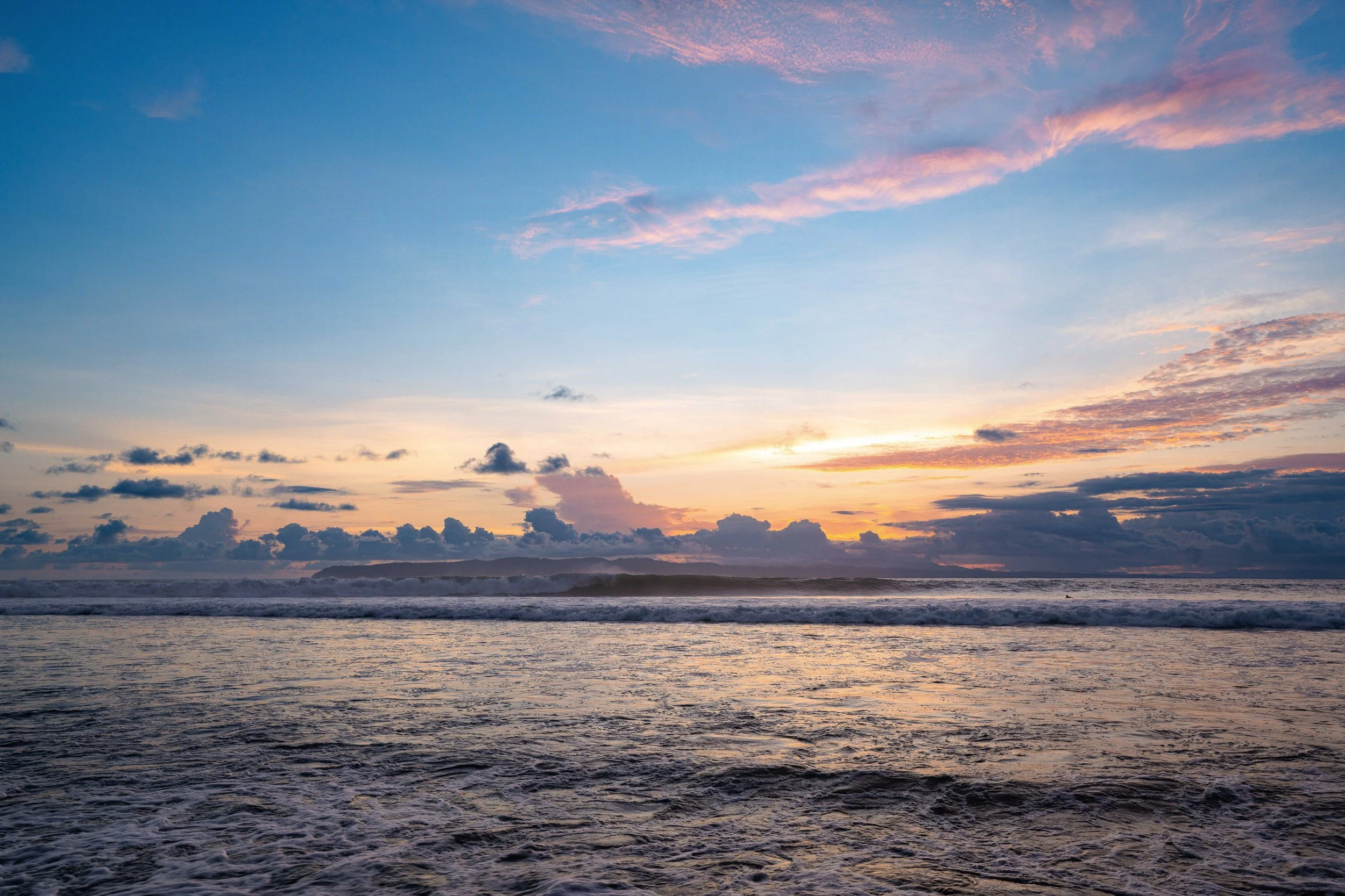 Scenic view of a sunset over the ocean with a sky filled with blue, pink, and purple clouds, and waves crashing on the shore.