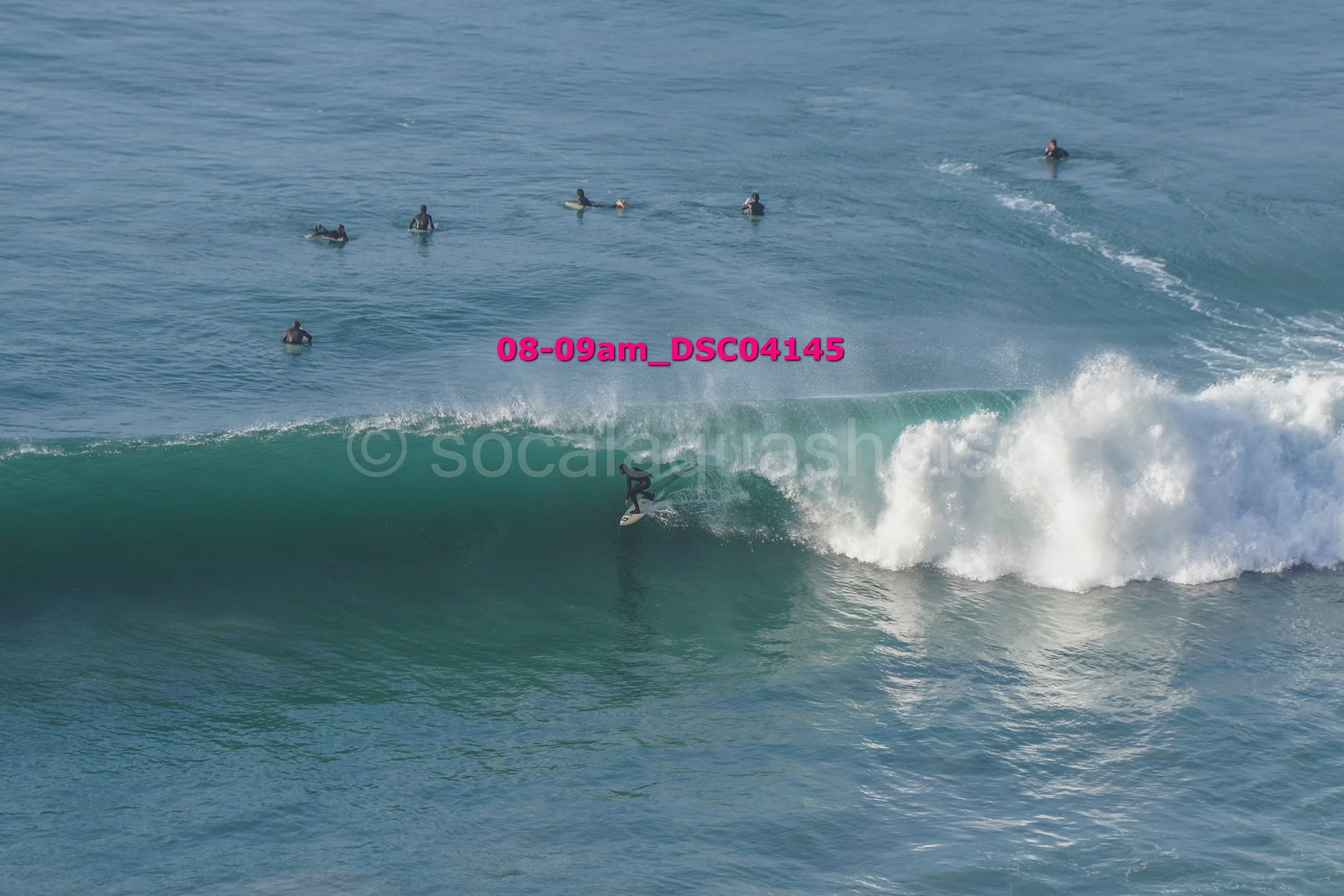 A person surfing on a large wave with several surfers floating in the water in the background.
