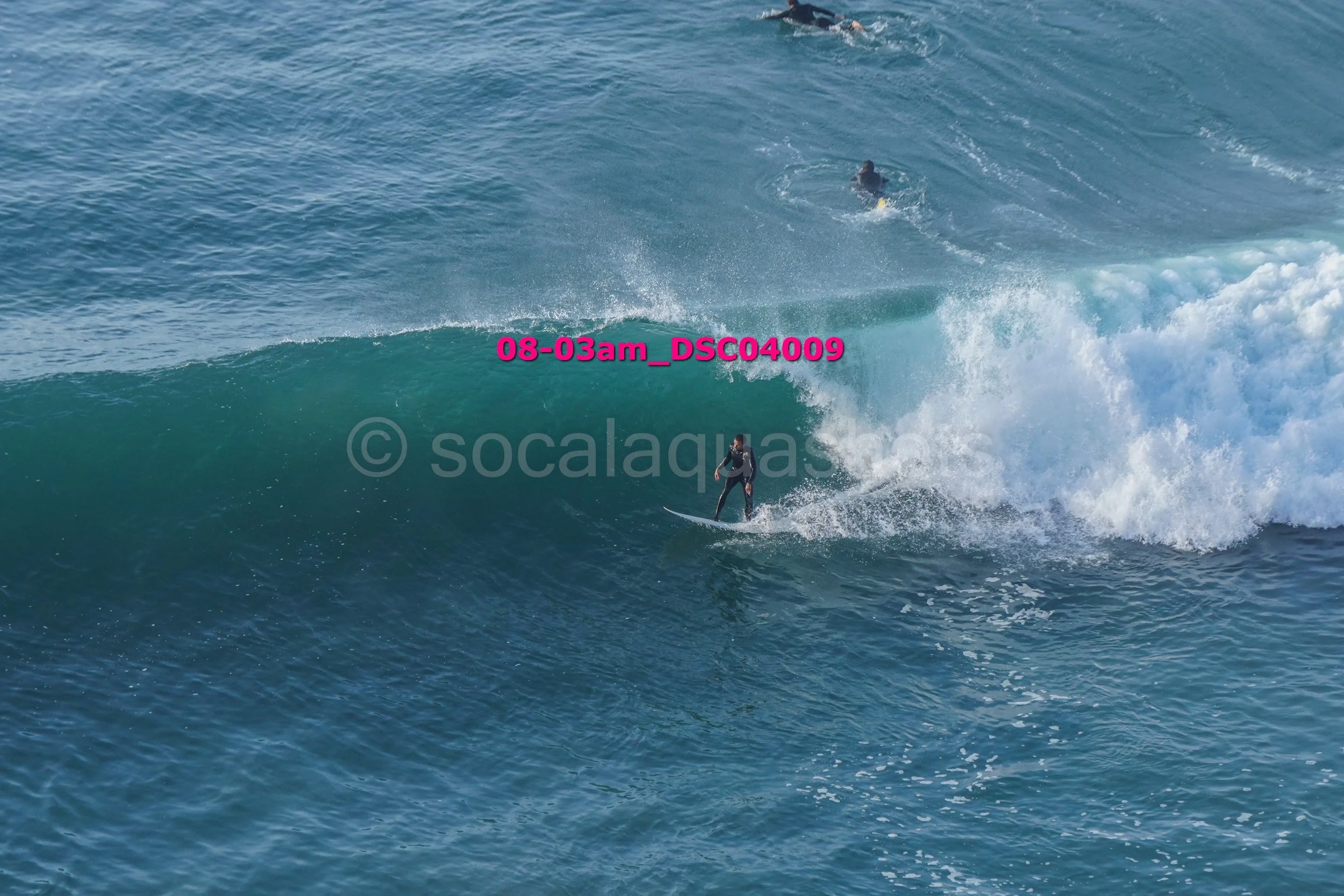 A person surfing on a large wave, with two other surfers visible in the background in the ocean.