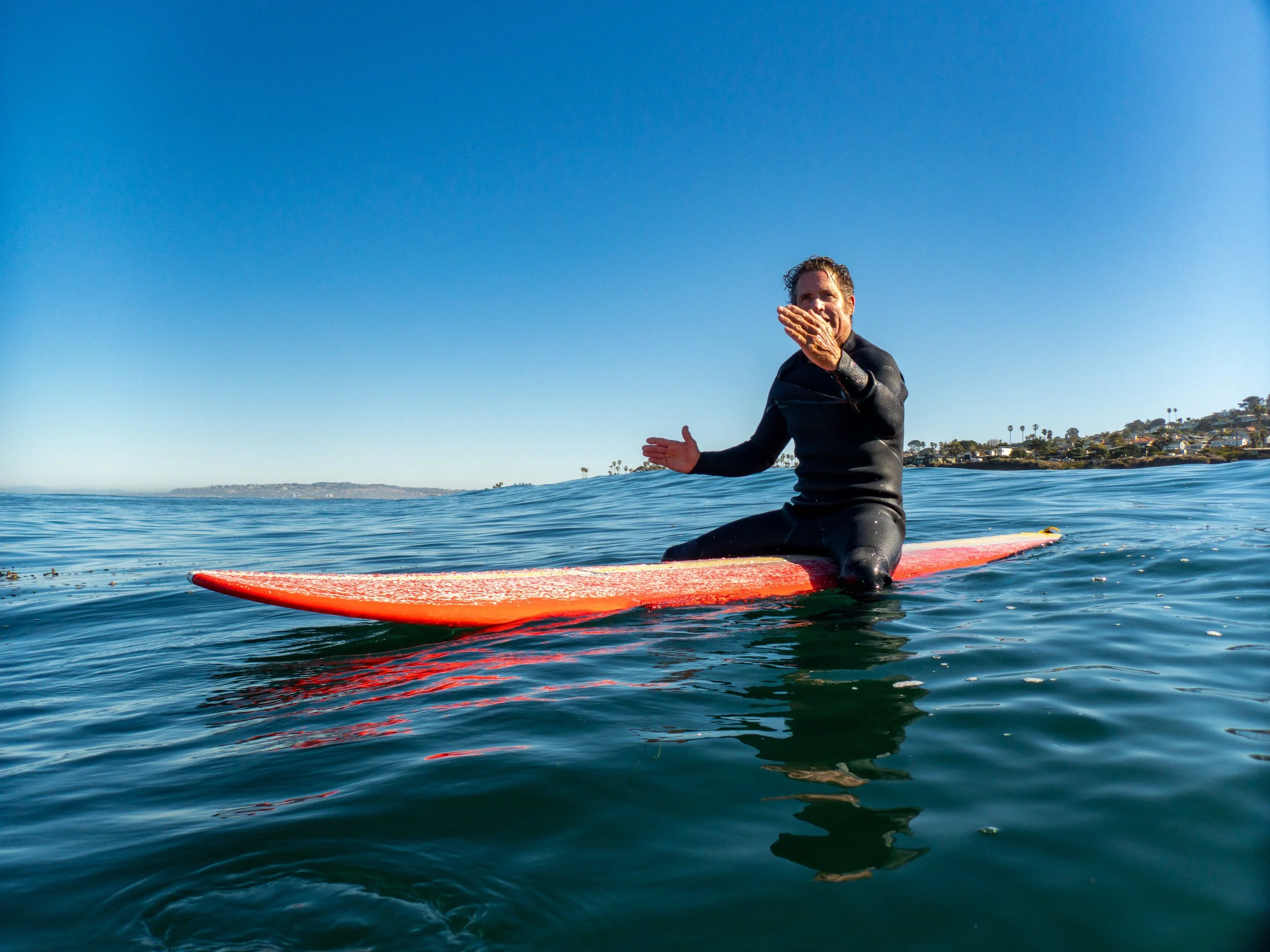 A man in a black wetsuit sitting on a red paddleboard in the ocean, smiling and waving, with a clear blue sky above and houses with palm trees along the shoreline in the background.