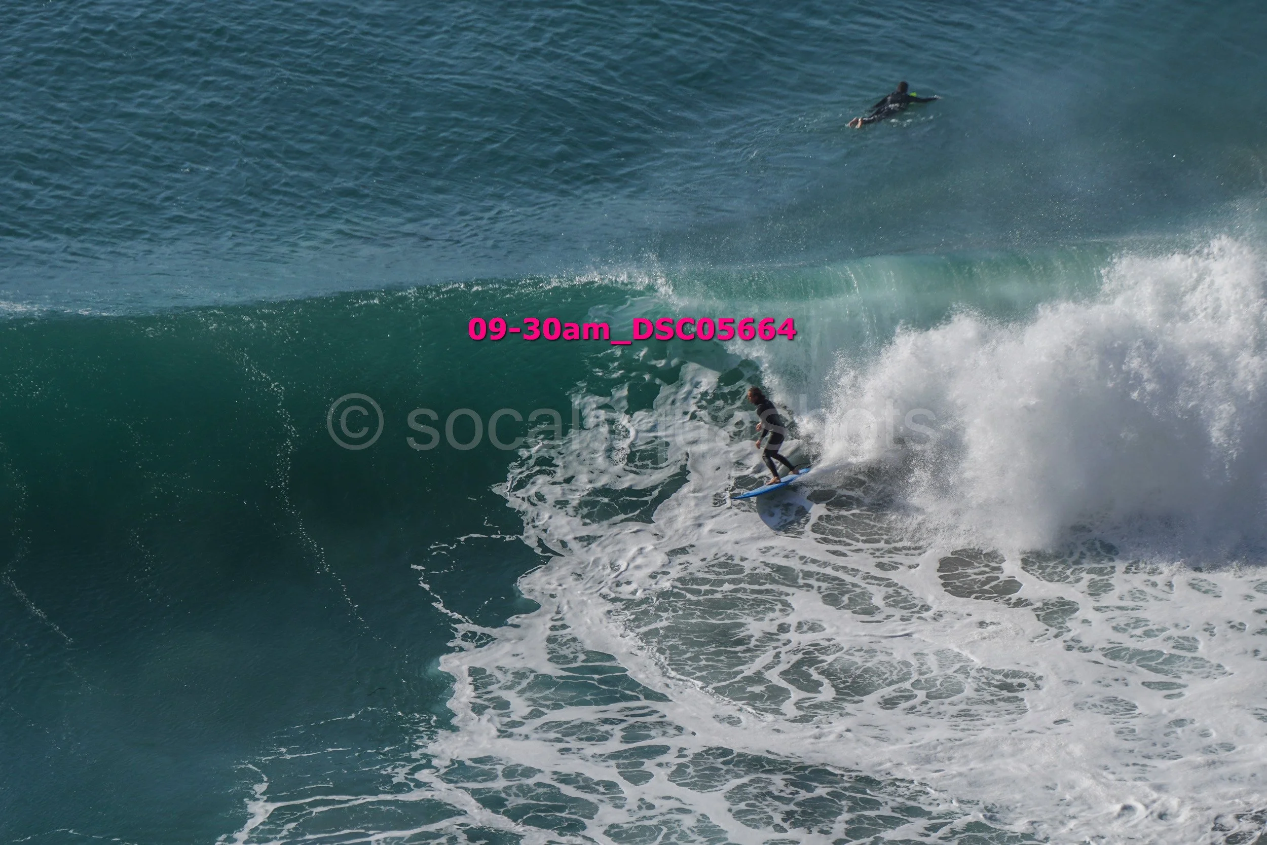 Surfing on a large wave with one surfer riding inside the wave and another surfer swimming in the water nearby.