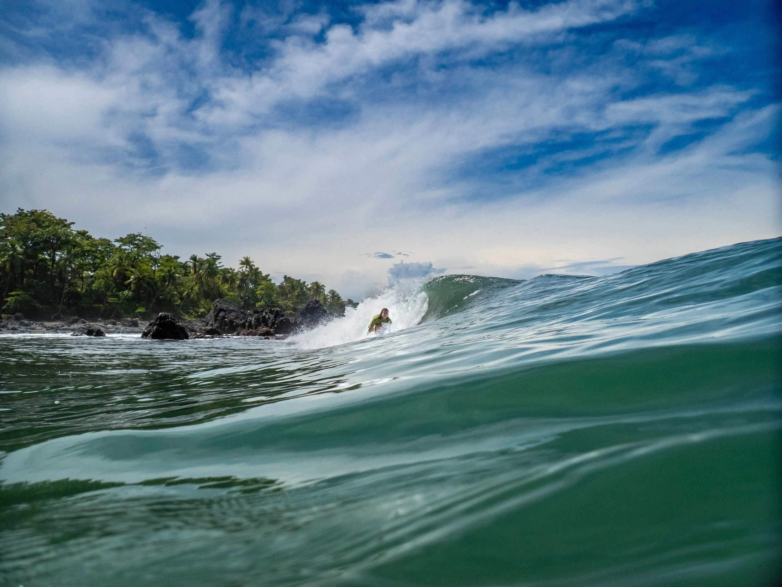 A person surfing on a wave near a rocky shoreline with tropical trees under a partly cloudy sky.