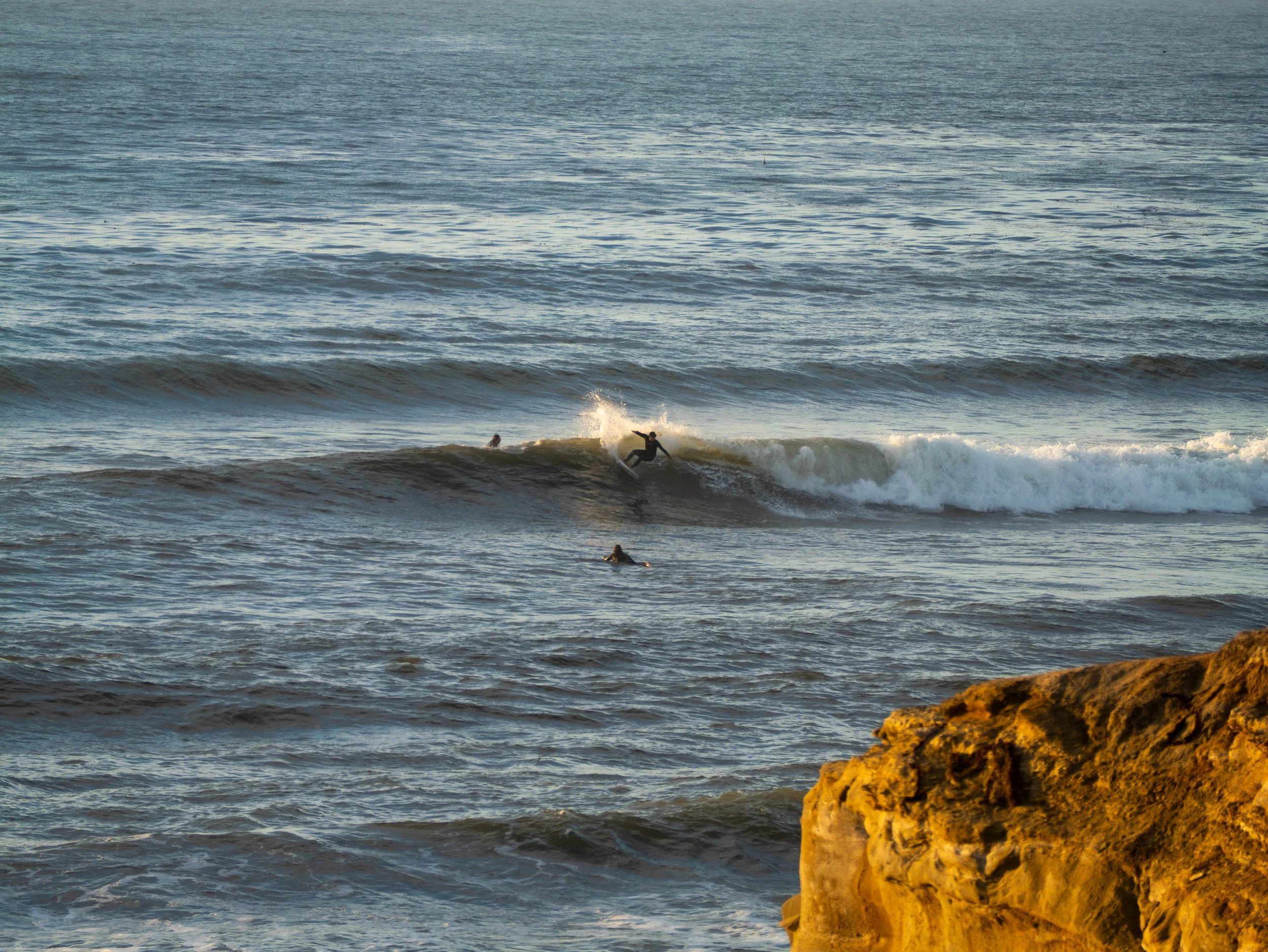 A person surfing on a wave with two other people in the water nearby, and a large rocky formation in the foreground.