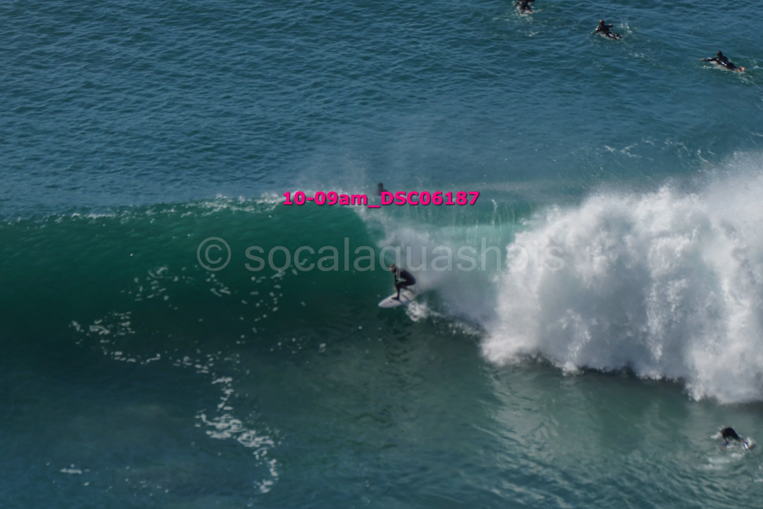 Surfer riding a wave in the ocean with several people swimming or surfing in the background.