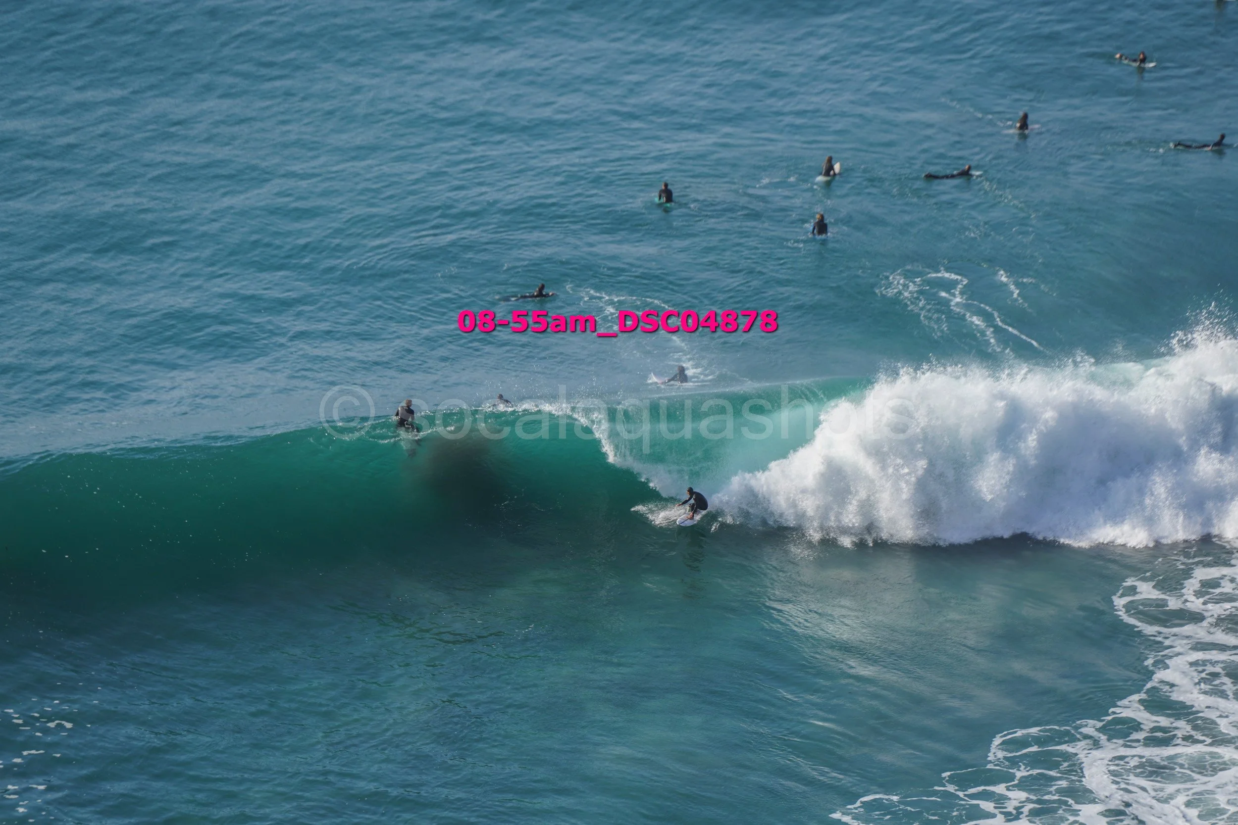 Surfer riding a large wave with multiple surfers in the water, some on surfboards and some swimming, in the ocean.