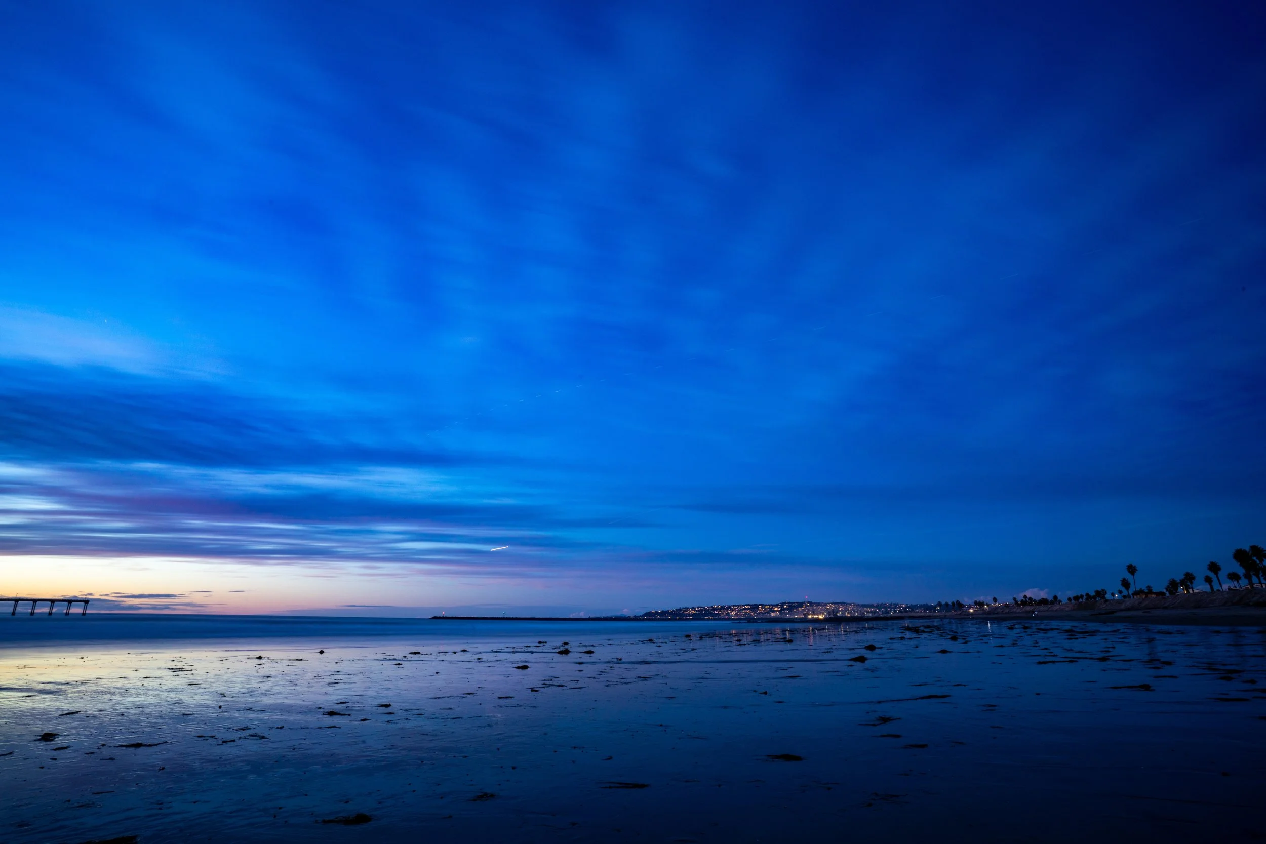 Calm ocean shoreline at dusk with a pier in the distance, a sky with streaks of clouds, and distant city lights on the horizon