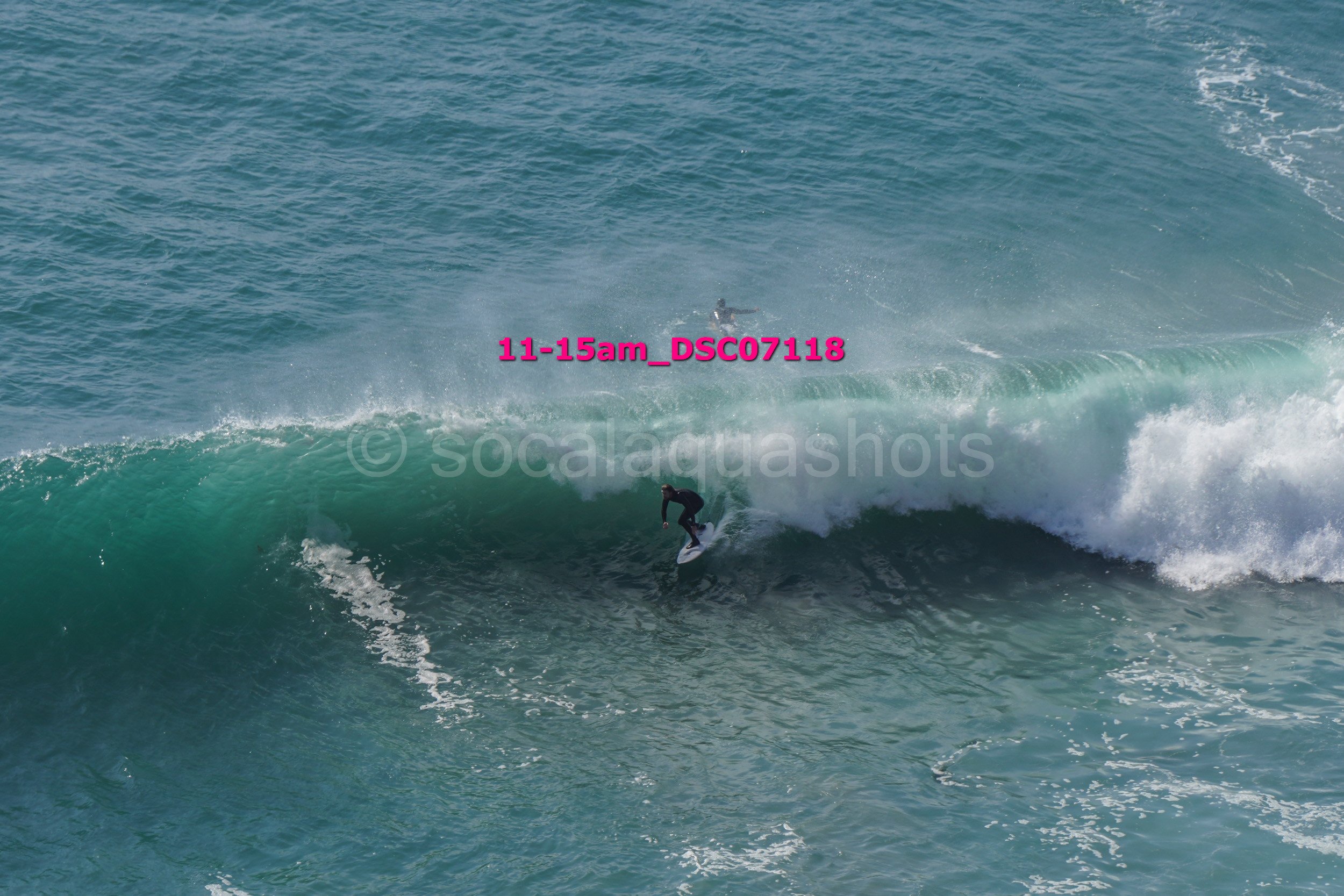 A surfer riding a large wave in the ocean, with another surfer visible in the background.