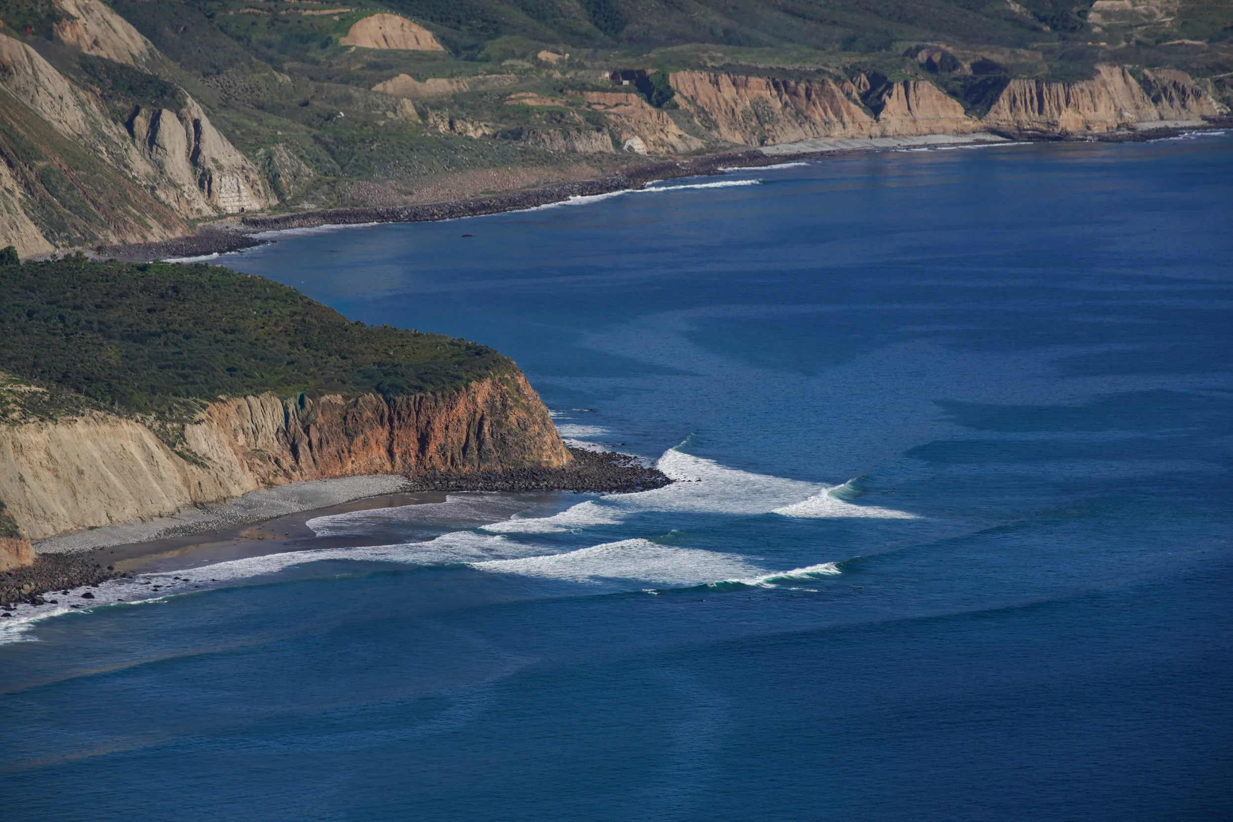 Coastal cliffs with waves crashing onto a pebble beach, overlooking a deep blue ocean.