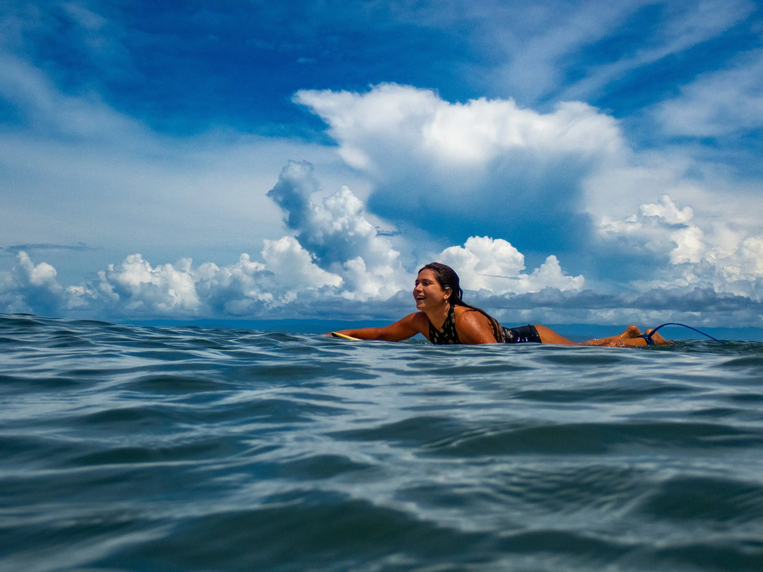 A woman in a swimsuit lying on a surfboard in the ocean, smiling with a backdrop of a partly cloudy sky.