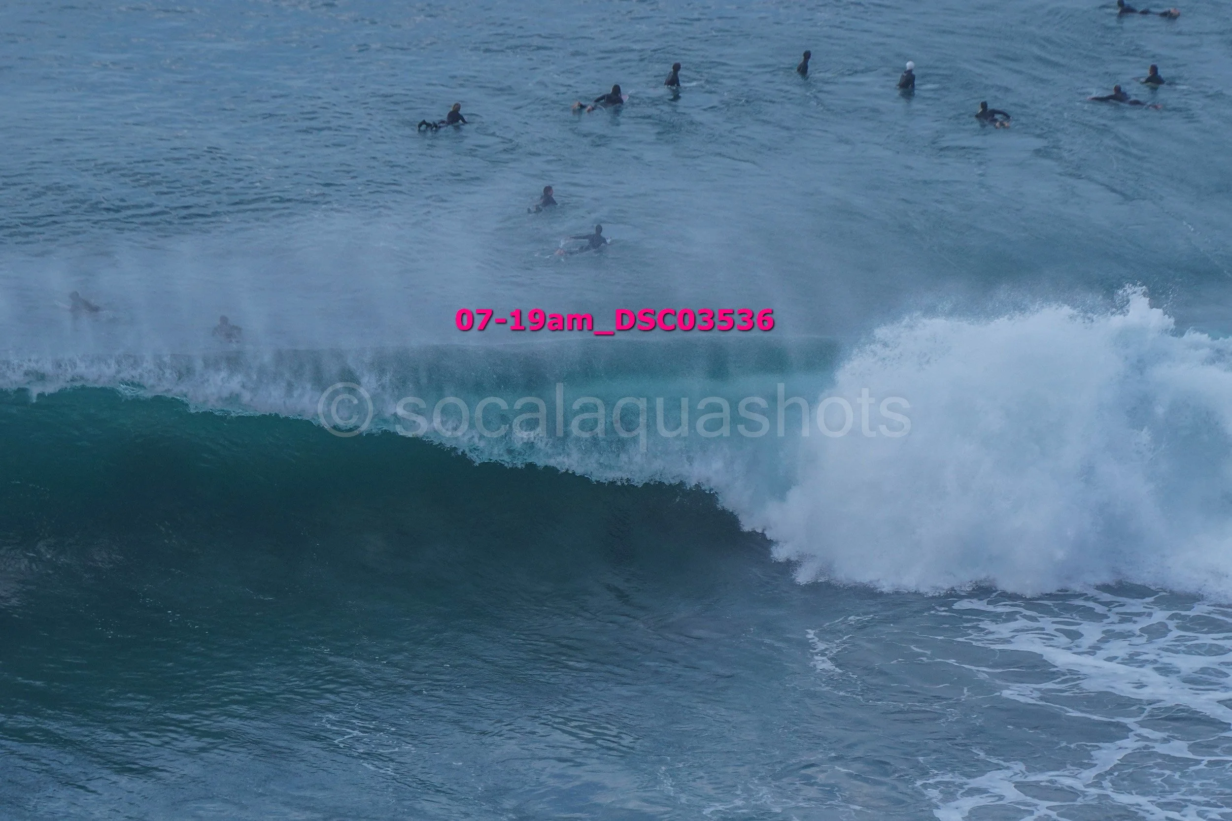 A group of surfers in the ocean on a wave, with some waiting and some riding, taken mid-morning during daylight.