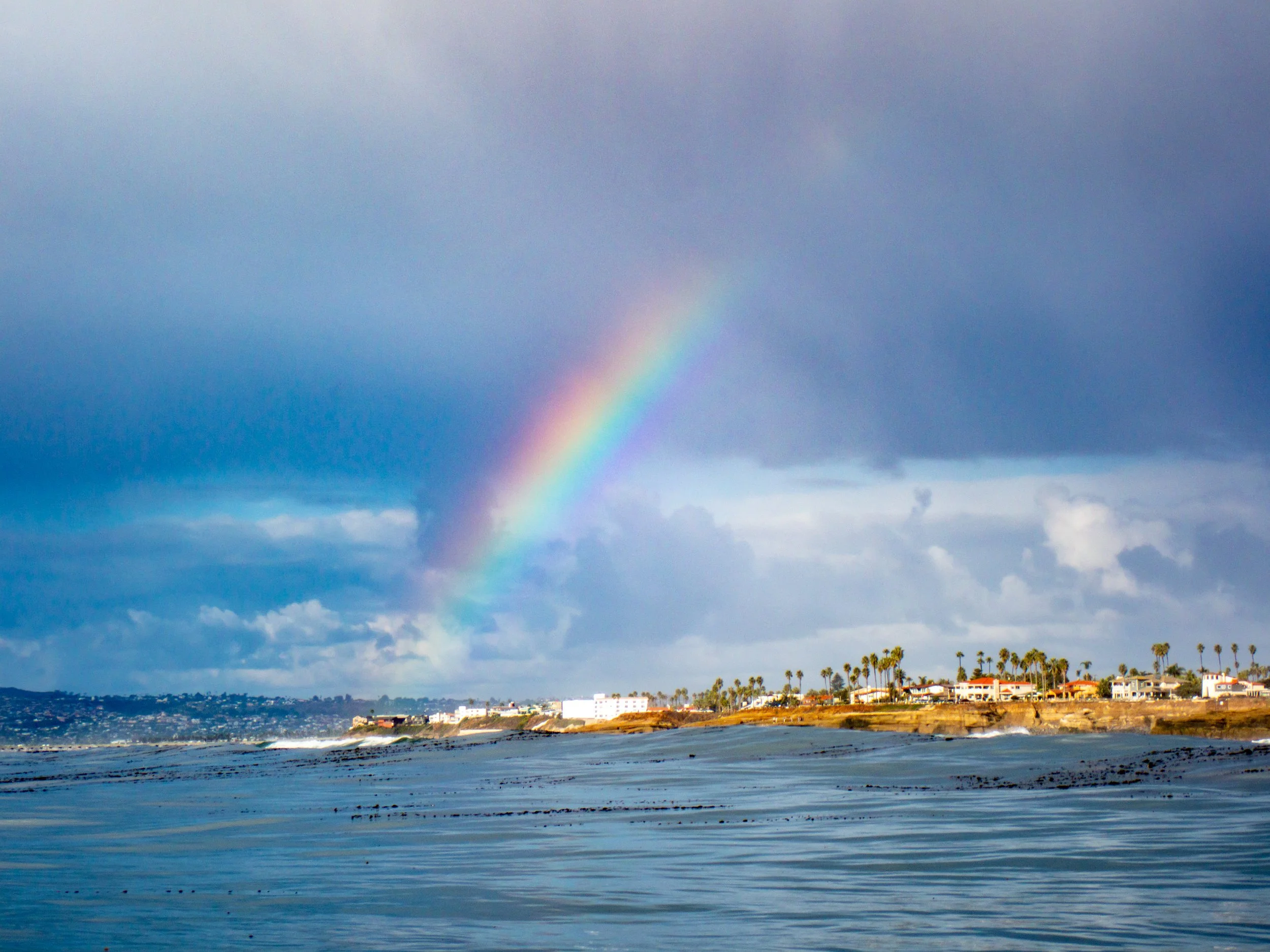 Rainbow over a beach coastal town with palm trees and houses on a cloudy day.
