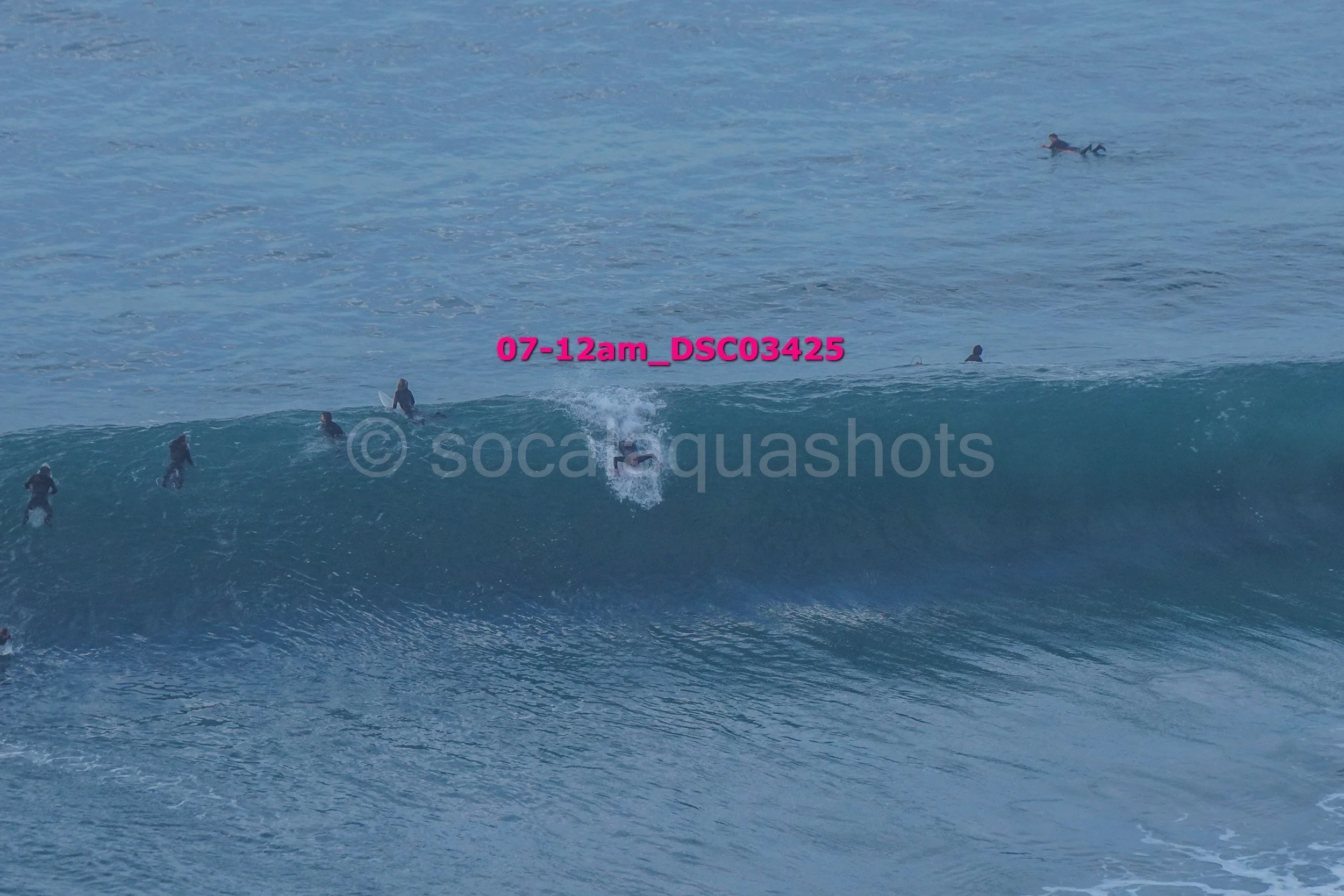 Surfer riding a large ocean wave with several surfers in the water around him, in a beach setting.