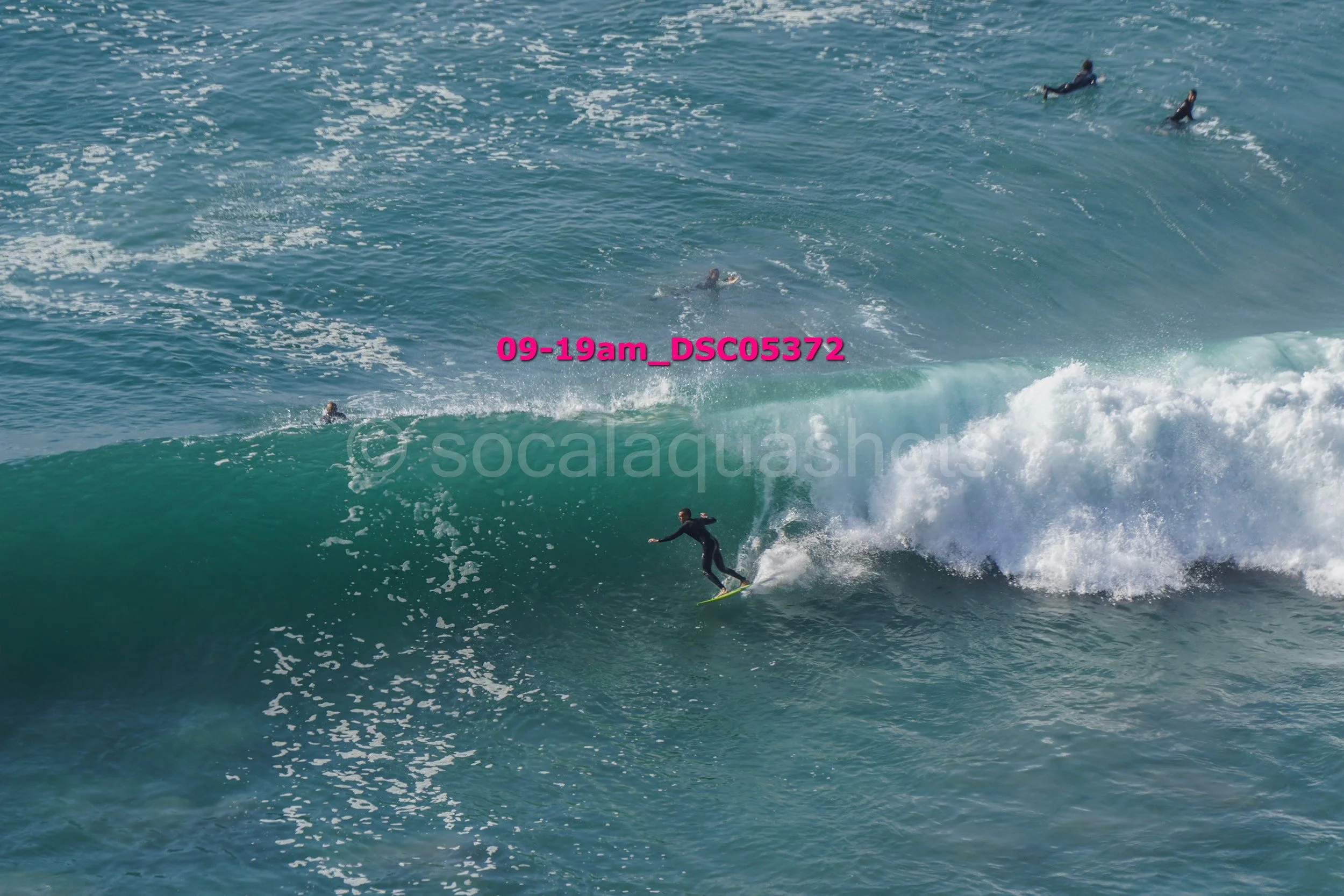 Surfer riding a large wave with several people swimming or surfing in the background.