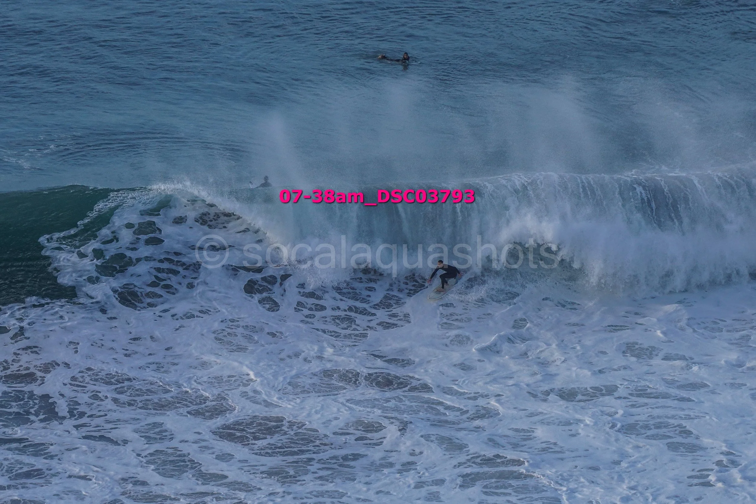 Surfer riding a large wave with two other surfers in the water behind, in an ocean setting.