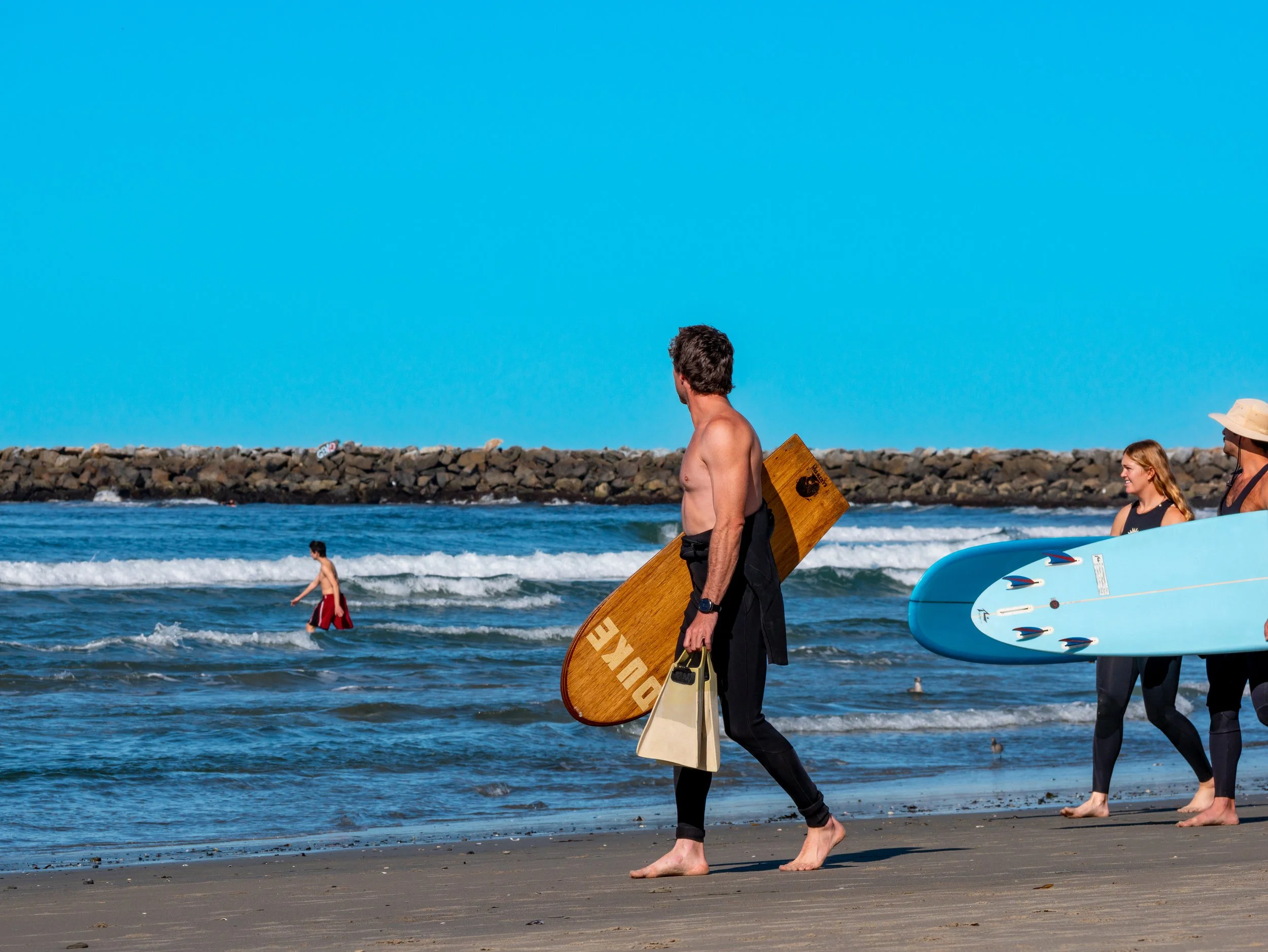 People walking on the beach with surfboards, a man carrying a wooden surfboard and a bag, two women with a blue surfboard, and a boy in the water, with a rocky breakwater in the background under a clear blue sky.