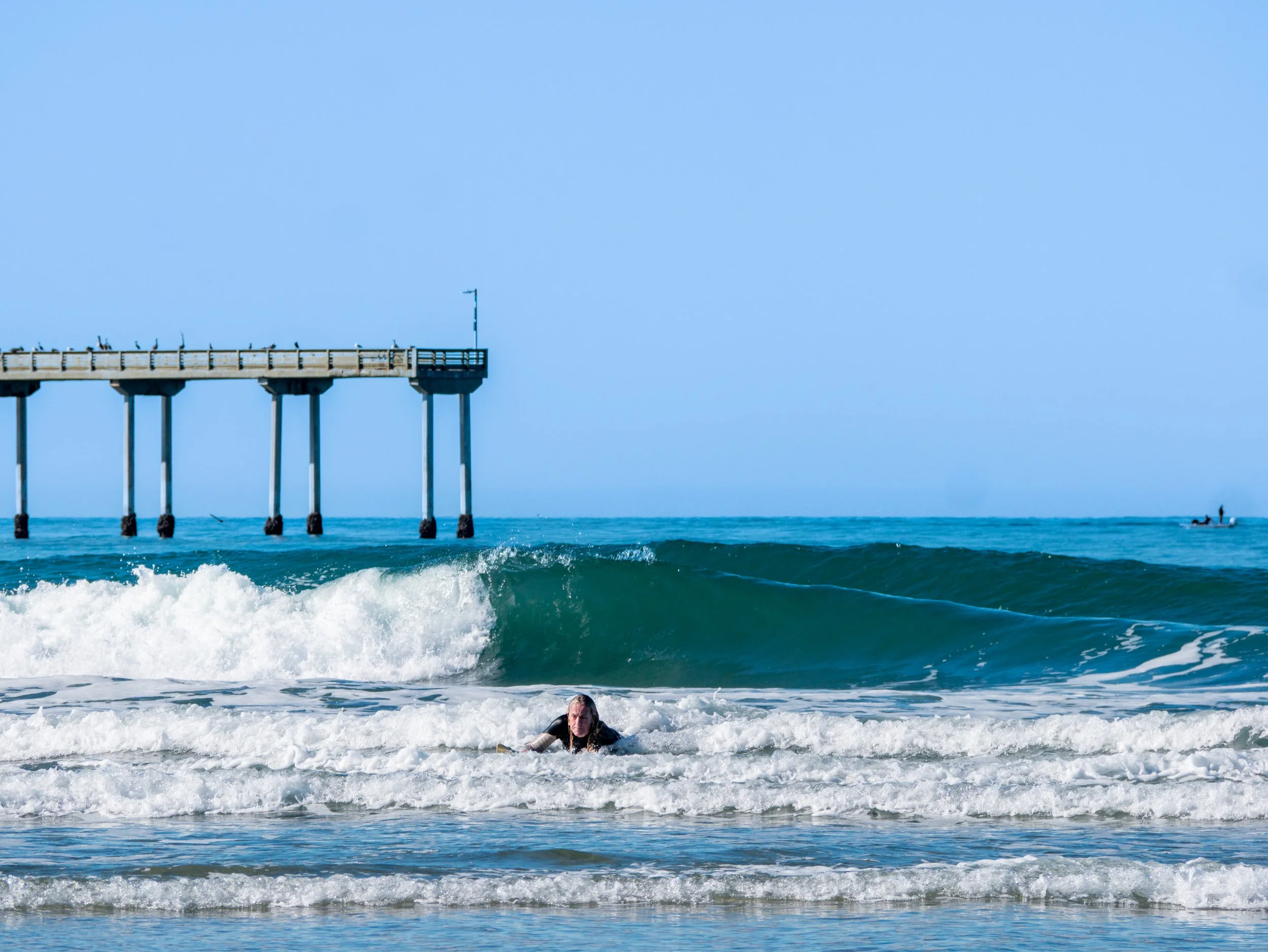 A person in the ocean close to the shore, lying on a surfboard, with a wave approaching. In the background, there is a pier extending into the water with a flag on it and a small boat further out in the sea.