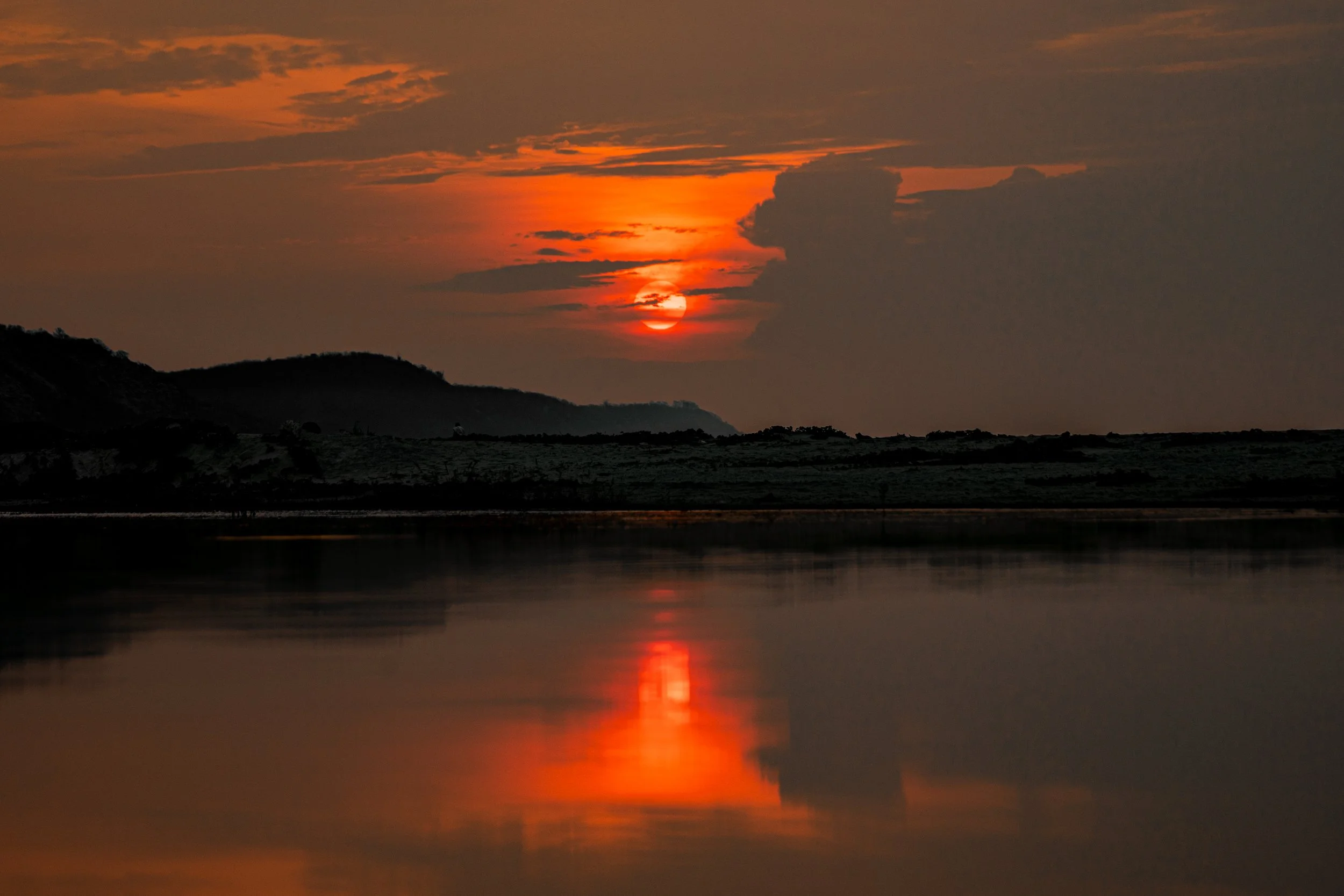 Sun setting over hills with orange and red hues, reflecting on a calm body of water in the foreground.