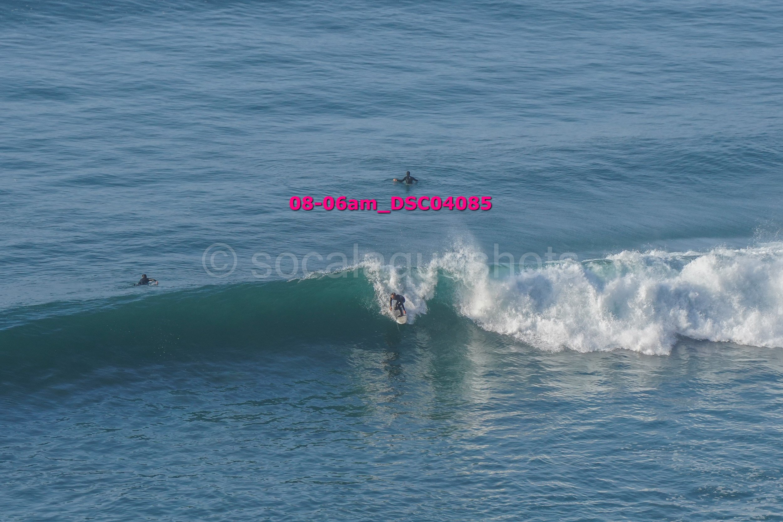 Surfer riding a wave with two surfers in the water nearby, ocean in the background.