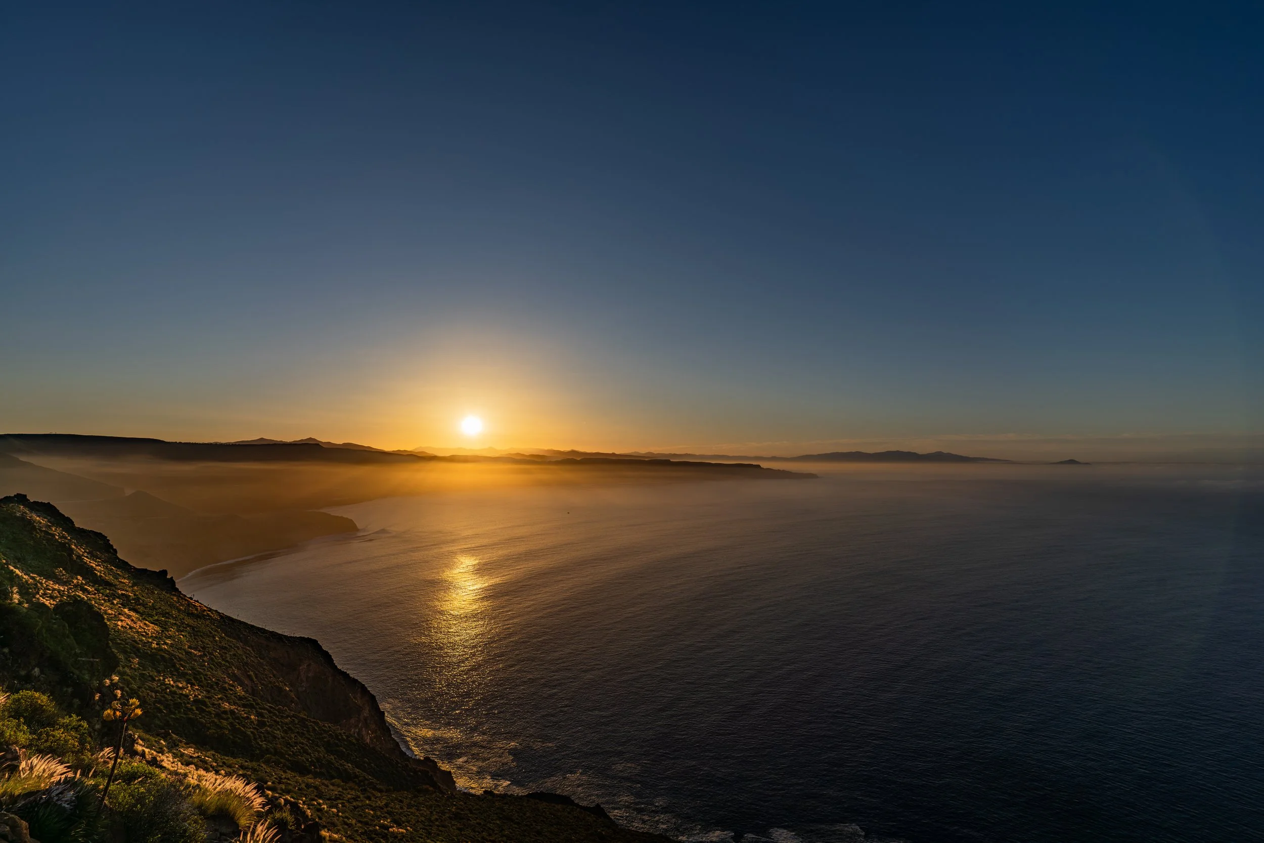 A sunset over a calm ocean with a hillside on the left and distant landforms on the horizon.