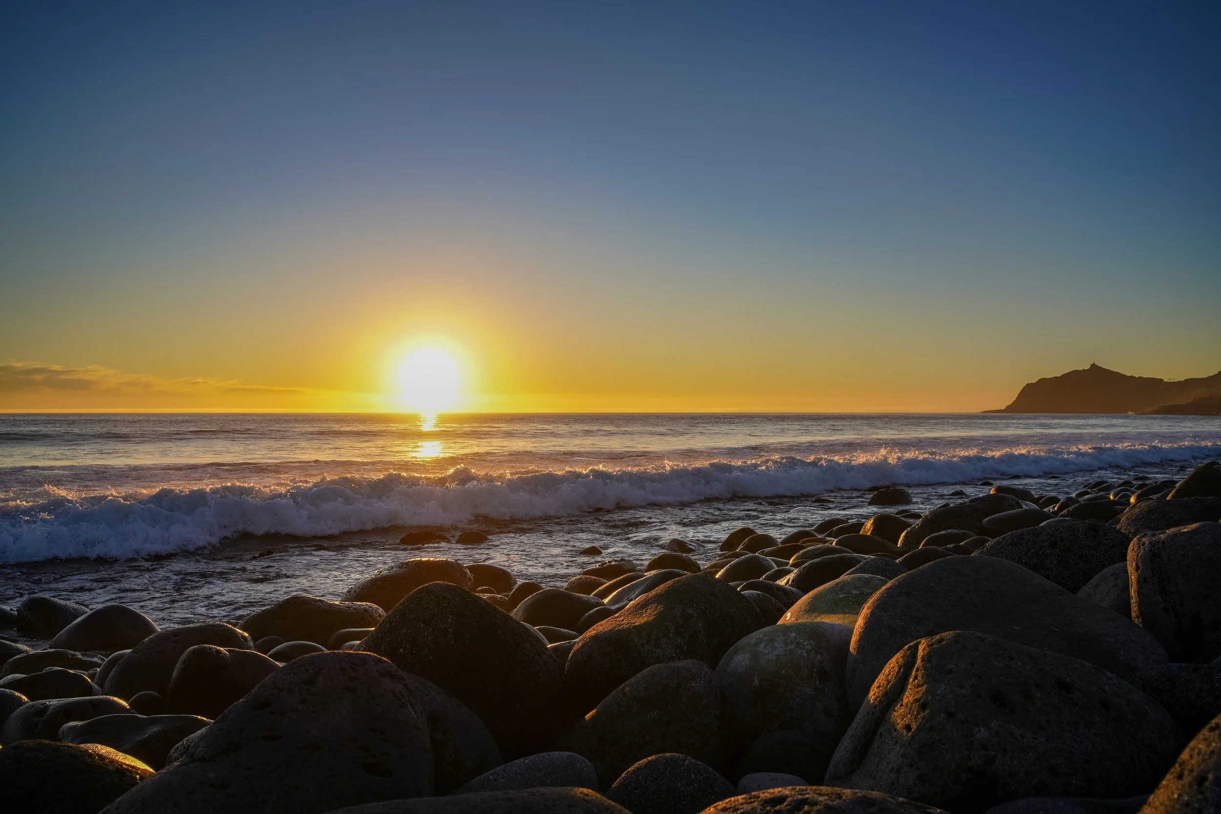 Sunset over the ocean with waves crashing onto a rocky beach and a distant hill on the right.