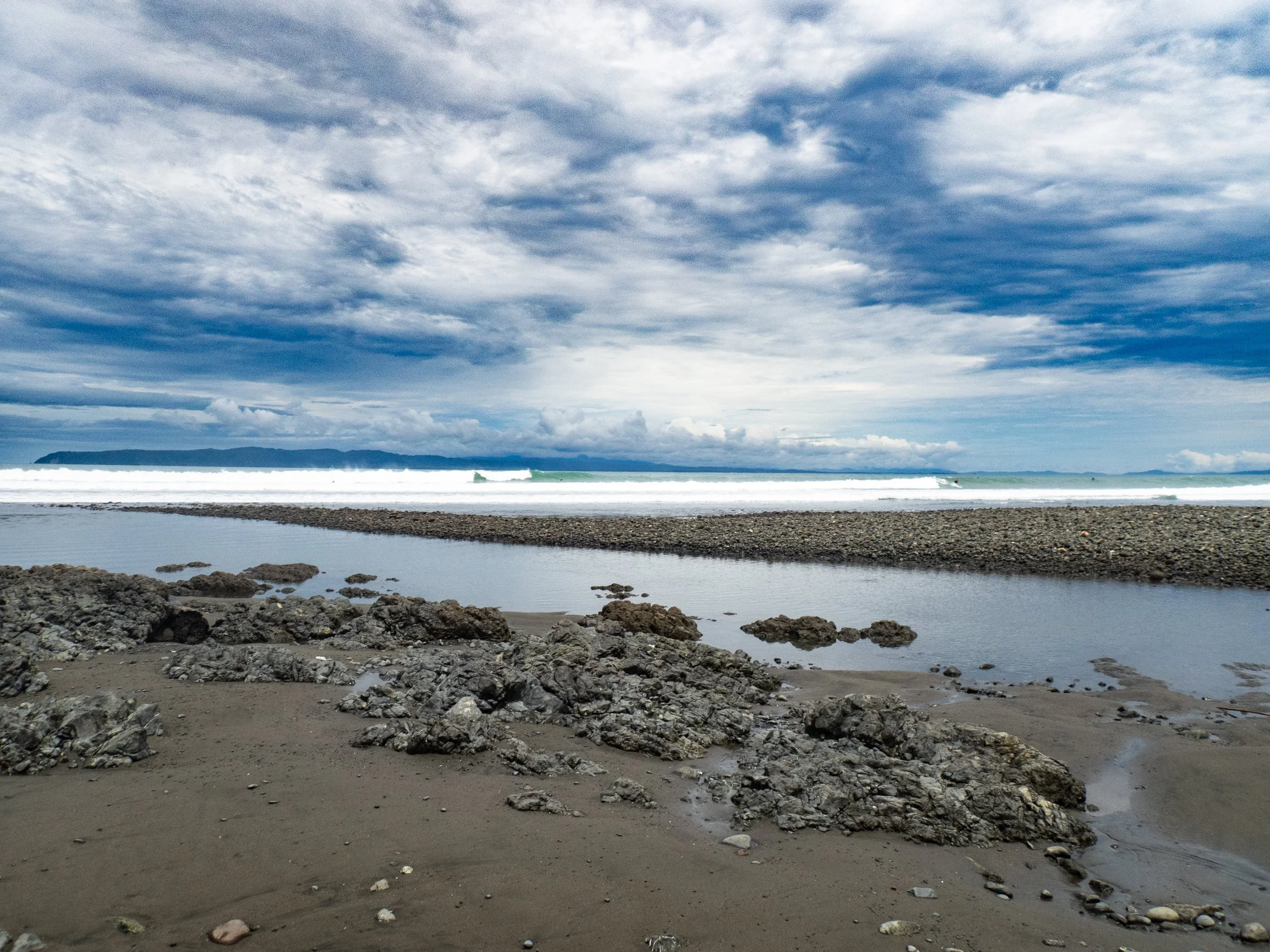A beach scene with cloudy sky, distant land on the horizon, rocks and sand in the foreground, and ocean waves.