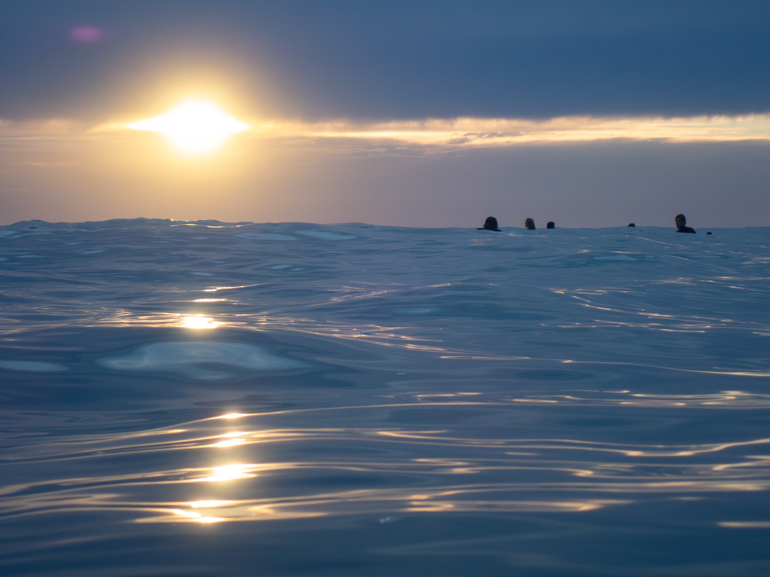 Ocean with gentle waves at sunset, silhouetted people partially submerged in water in the distance, and dark clouds above.