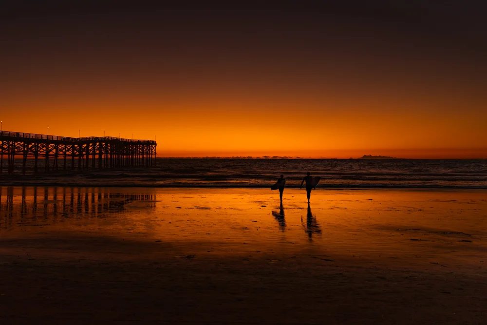 Two surfers silhouetted on wet sand at sunset with a pier and copper-colored reflections.