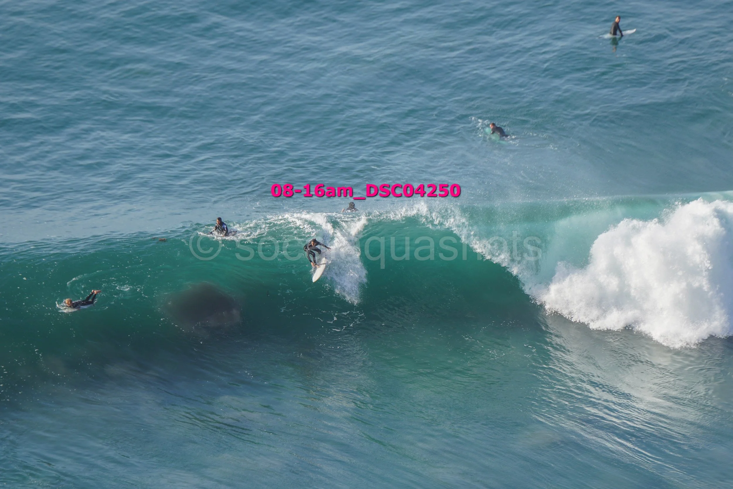 Surfer riding a large wave, with several other surfers in the water, at a beach.