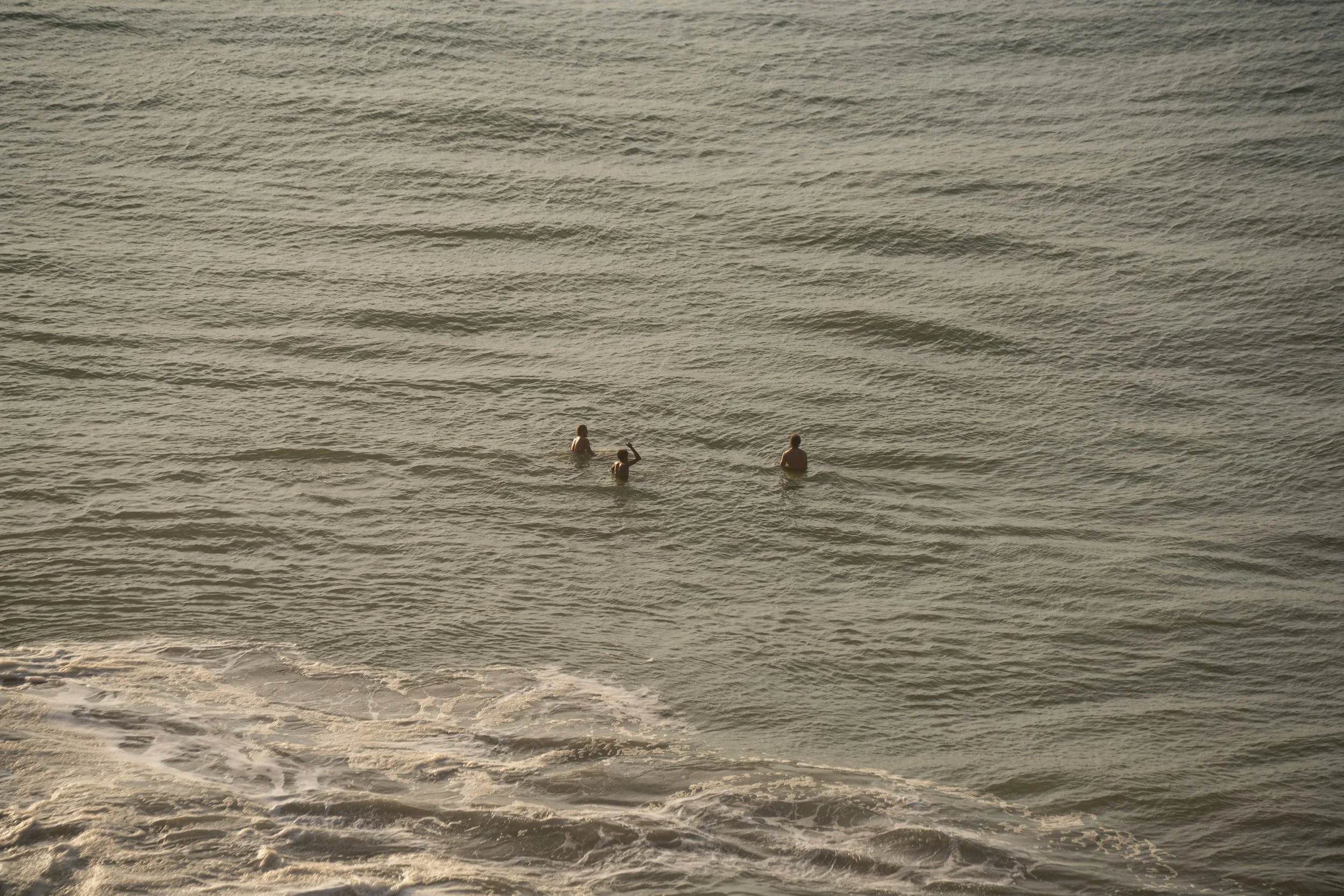 People swimming and wading in the ocean near the shoreline at sunset.