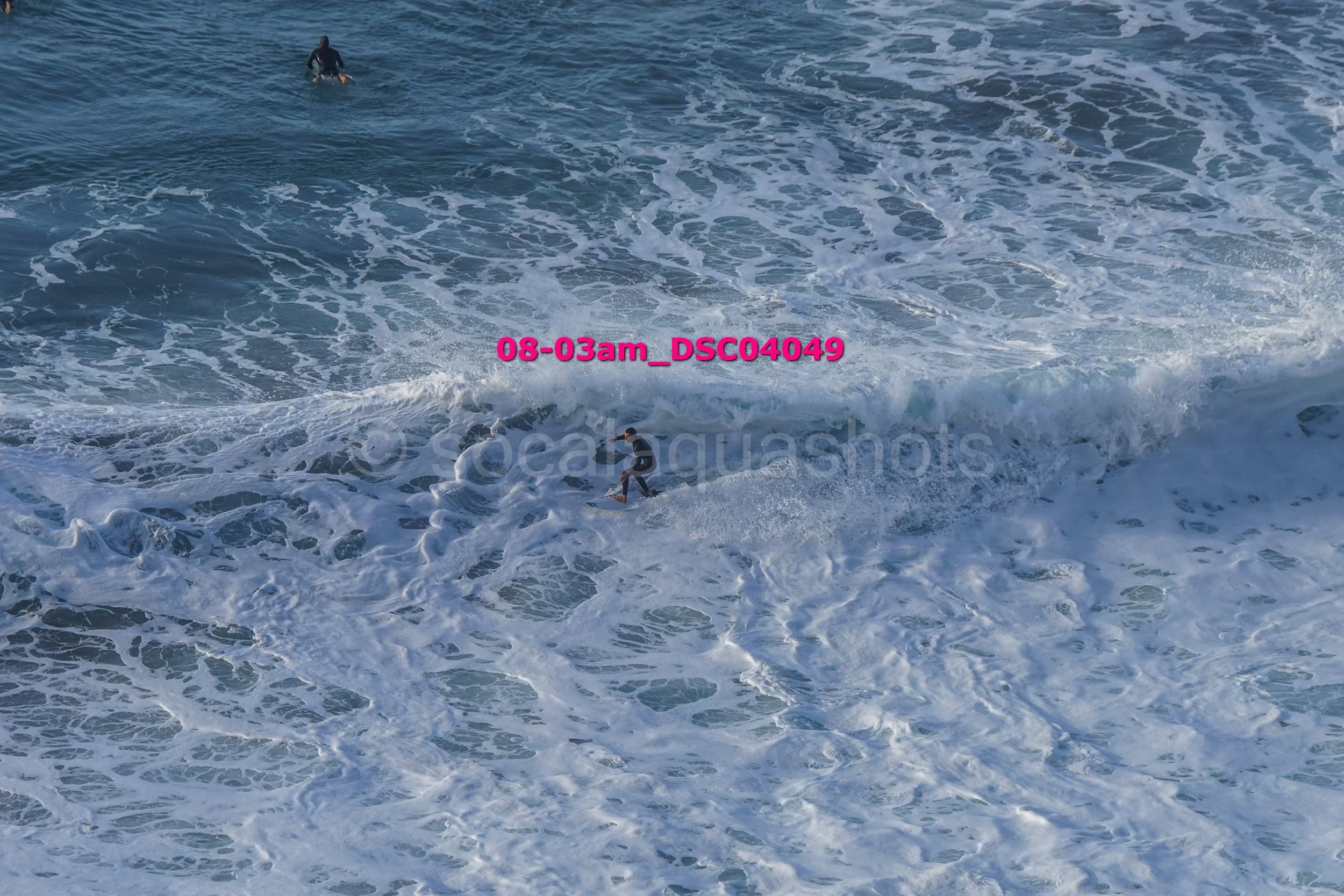 A person surfing on a wave in the ocean during daytime, with another person visible further in the water.