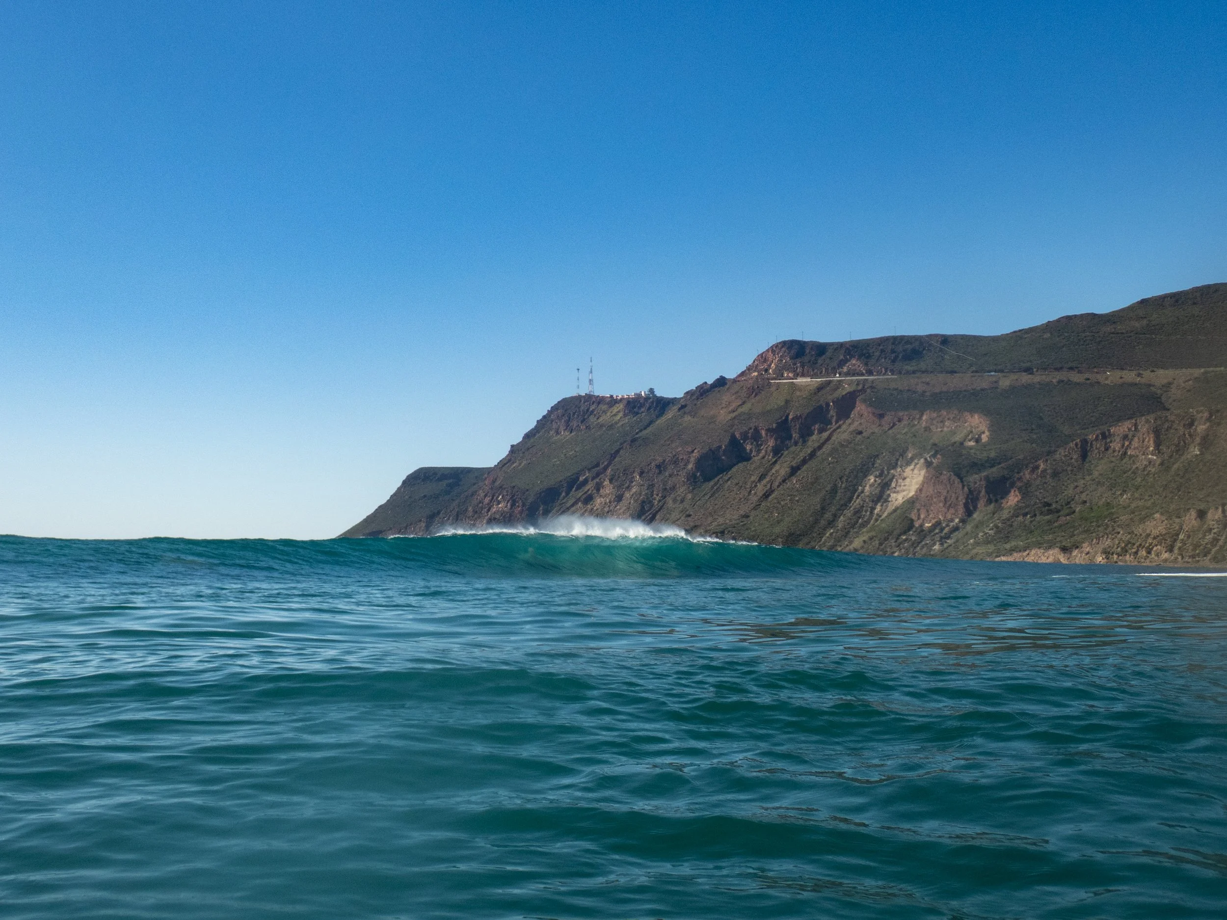 Ocean waves with a mountain coastline under a clear blue sky.