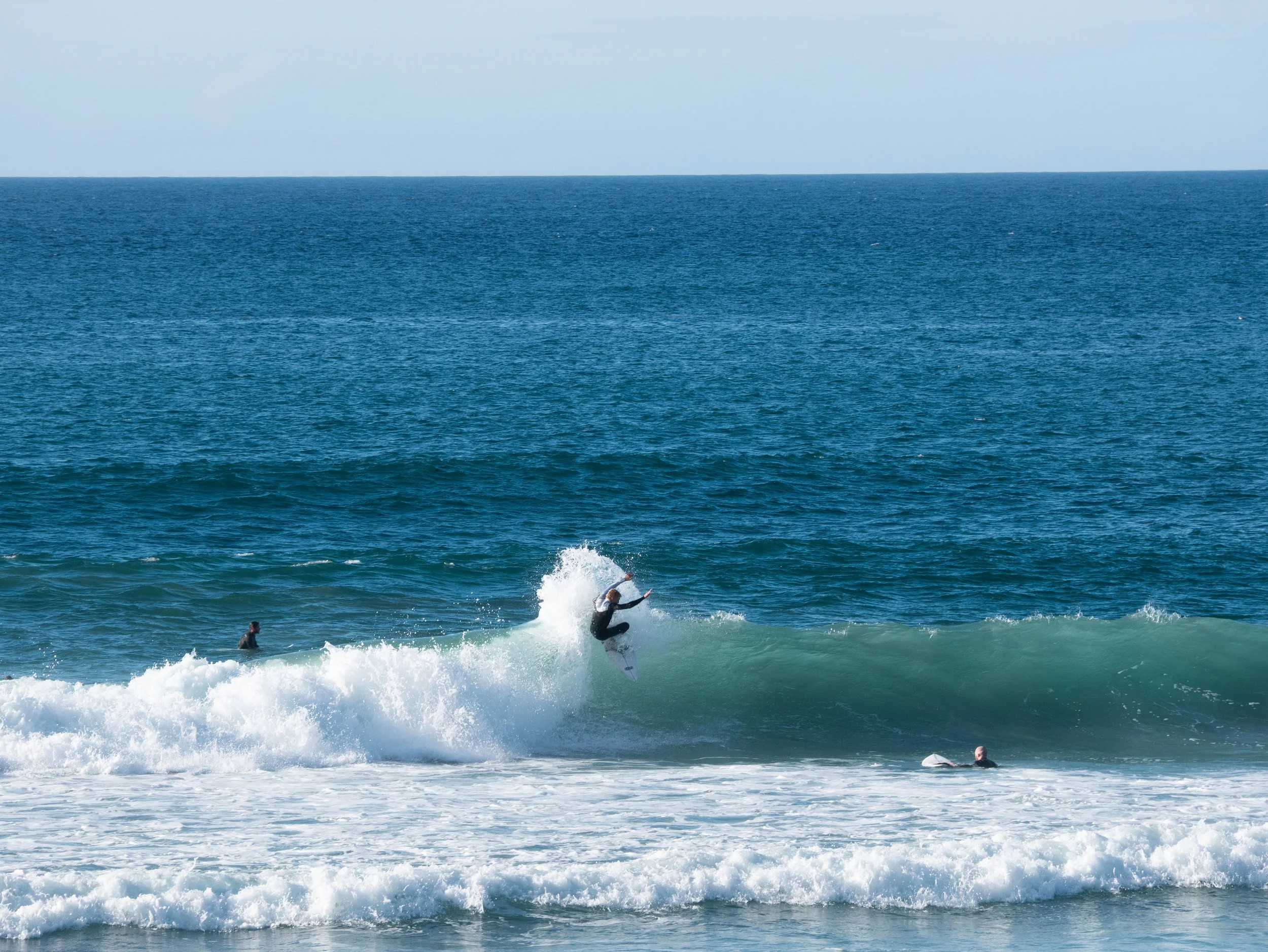 A person in a wetsuit surfing on a wave at the beach with other surfers nearby and the open ocean in the background.