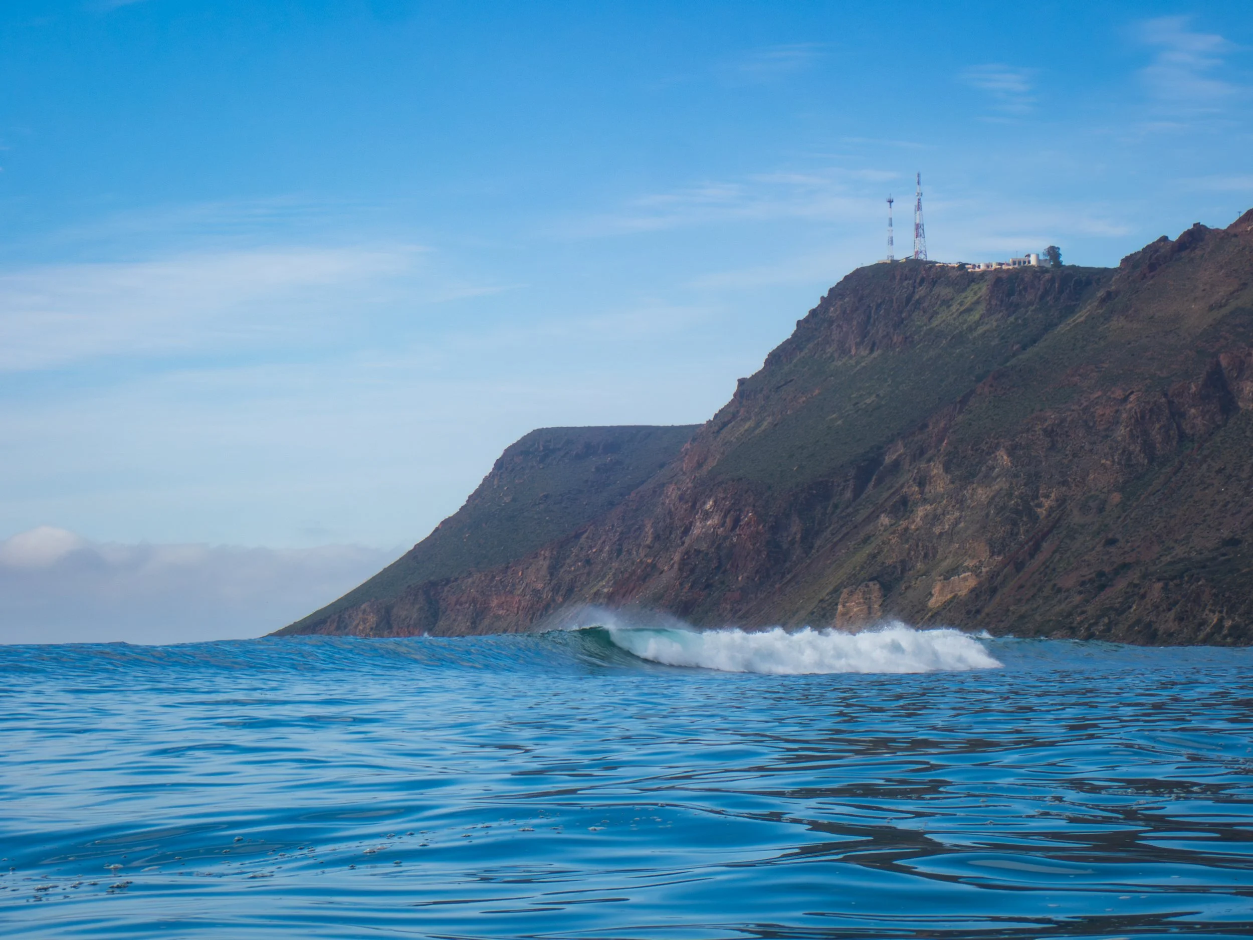 Ocean waves breaking near a rocky hillside with communication towers on top, under a blue sky with wispy clouds.