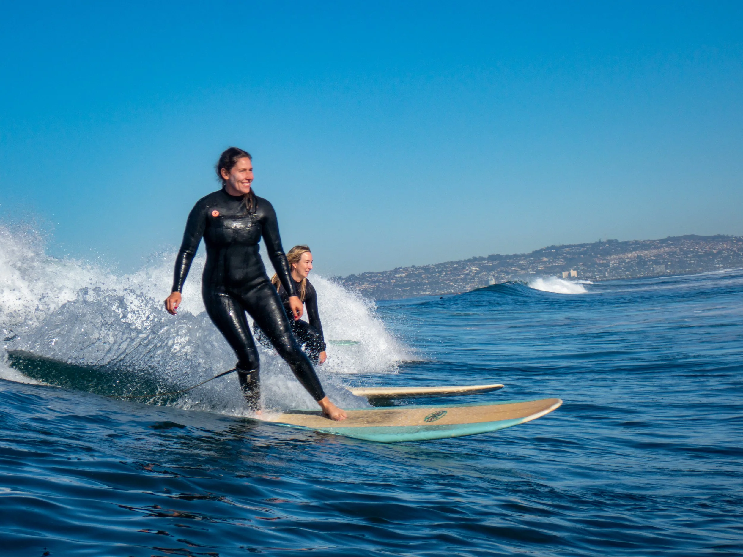 Two women in wetsuits stand on surfboards riding a wave in the ocean under a clear blue sky, with a distant shoreline visible in the background.