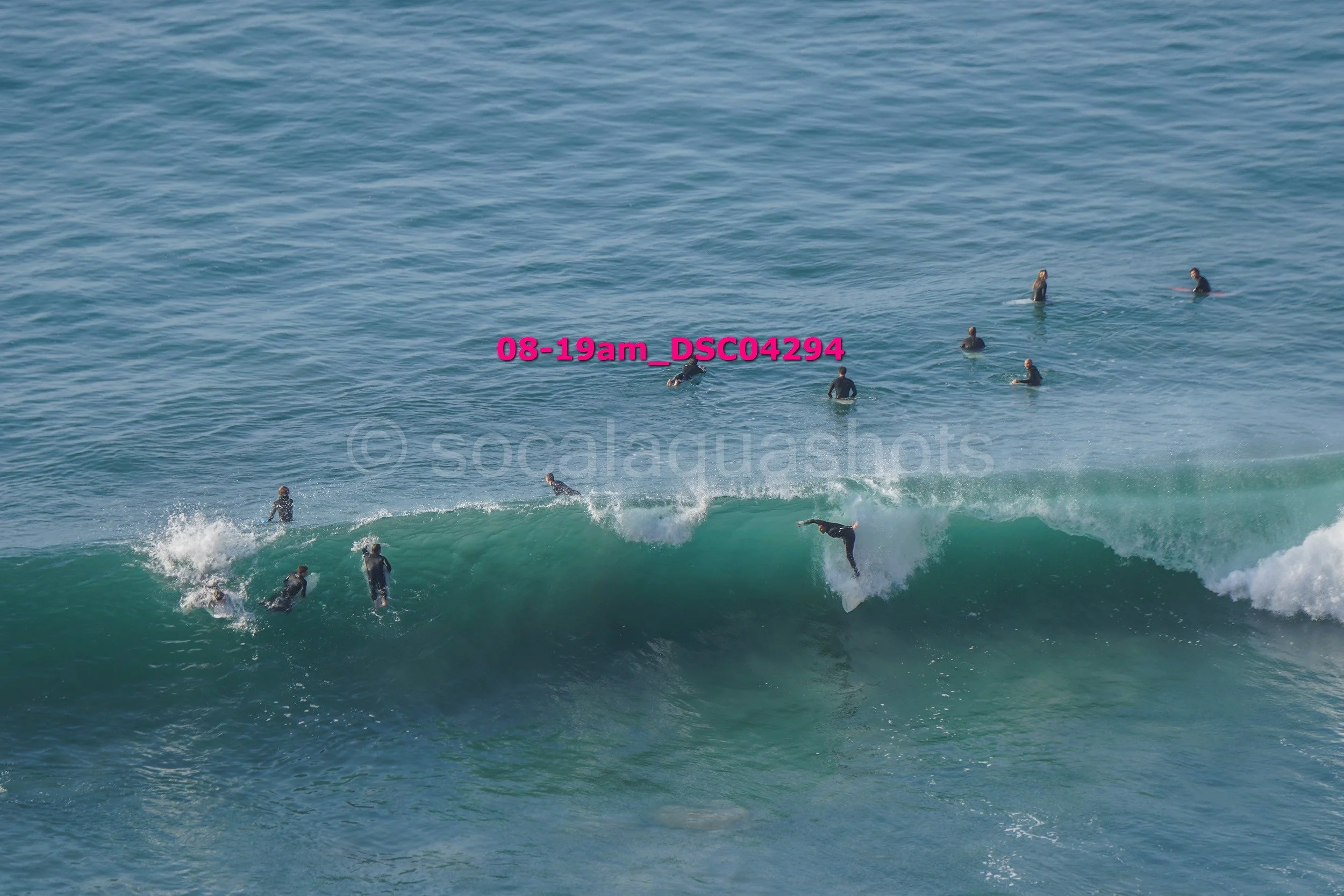 Surfers riding and practicing on large ocean waves with several people in the water, some standing and some surfing.