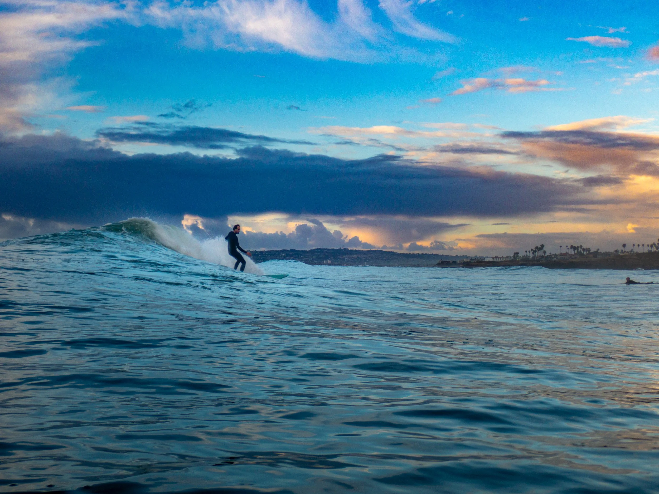 A person surfing on a wave in the ocean during sunset with clouds and distant shoreline in the background.