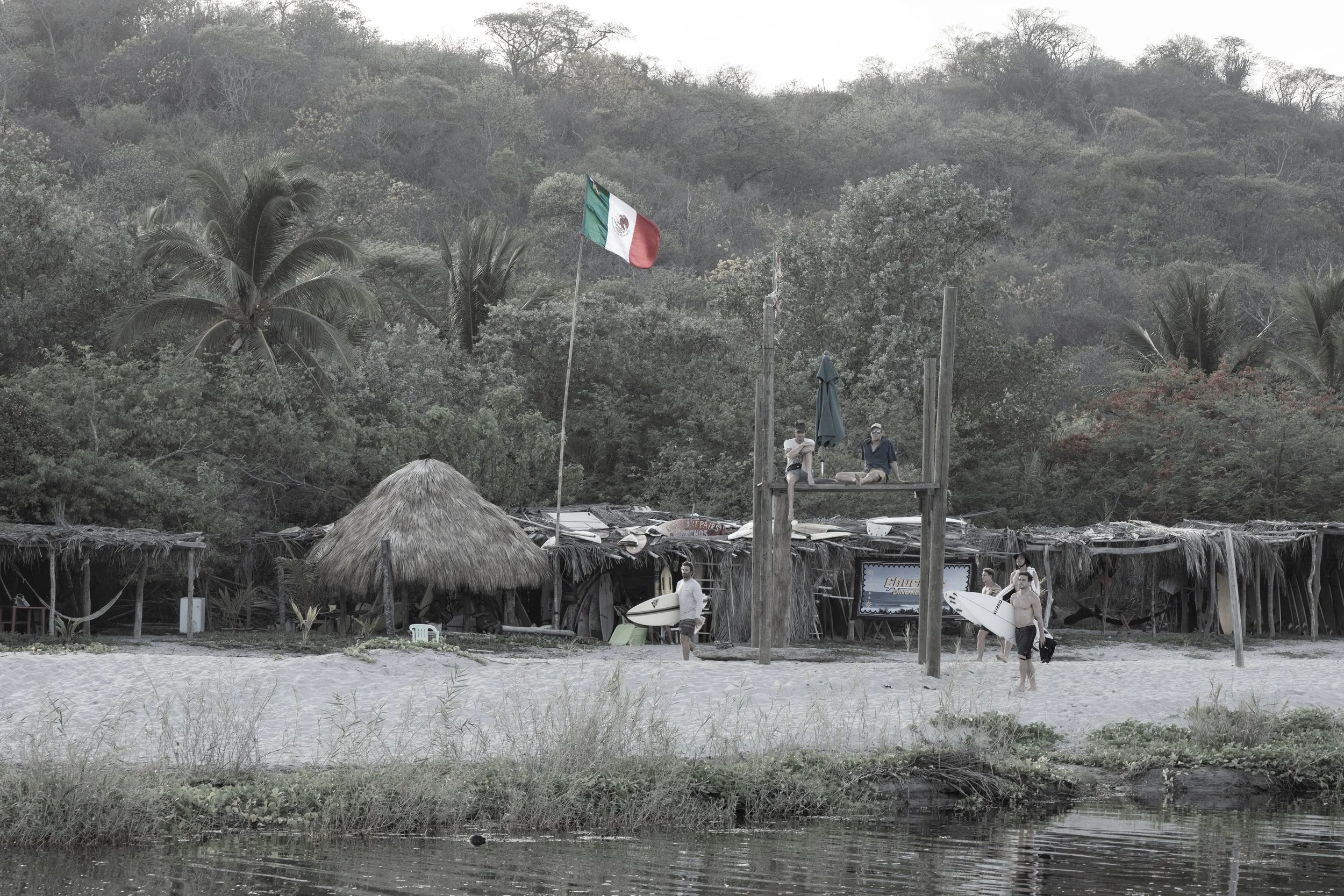 A beach scene with a thatched roof hut, people relaxing on a platform with a Mexican flag, surrounded by trees and a body of water in the foreground.