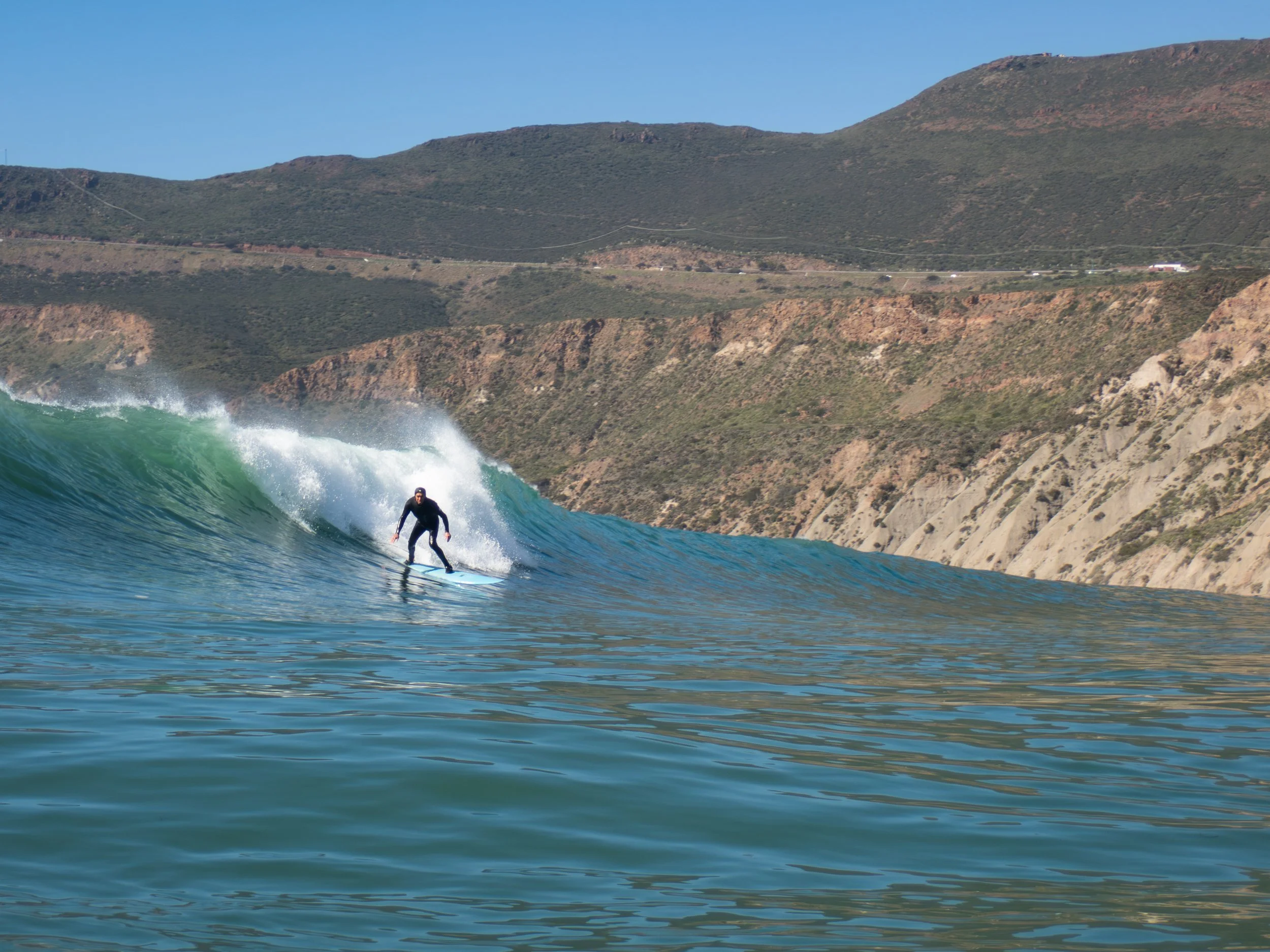 A person in black wetsuit surfing a wave near a rugged coastline with hills and sparse vegetation under a clear blue sky.
