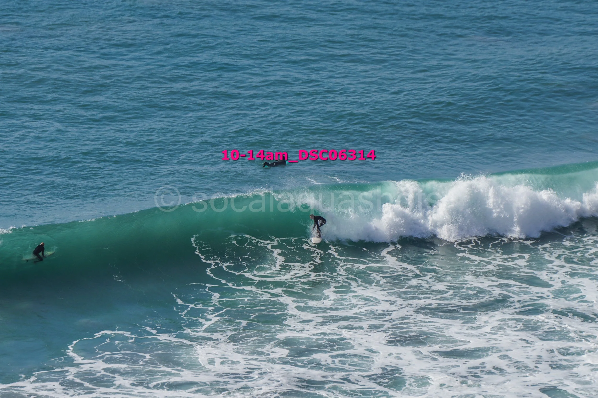 Three surfers riding and waiting for waves in the ocean, with one surfer mid-ride on a wave and two others paddling nearby.